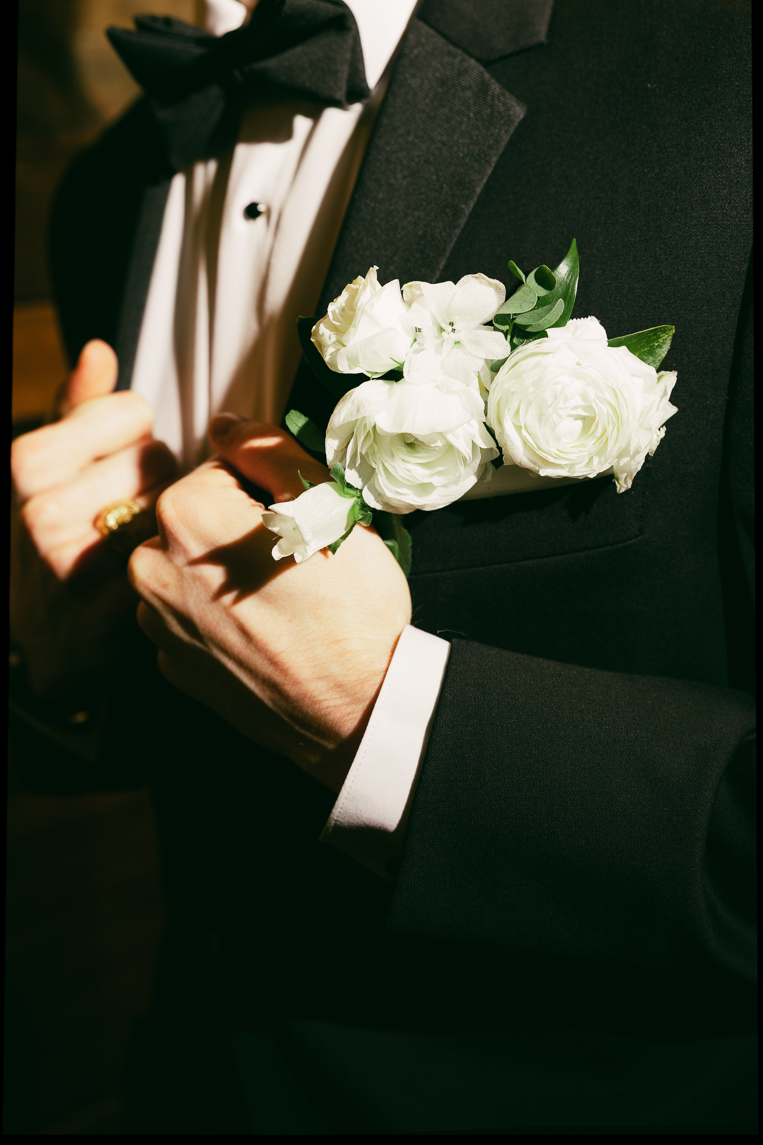 A man dressed in a tuxedo with a white dress shirt and black bow tie, holding a bouquet of white flowers over his chest.