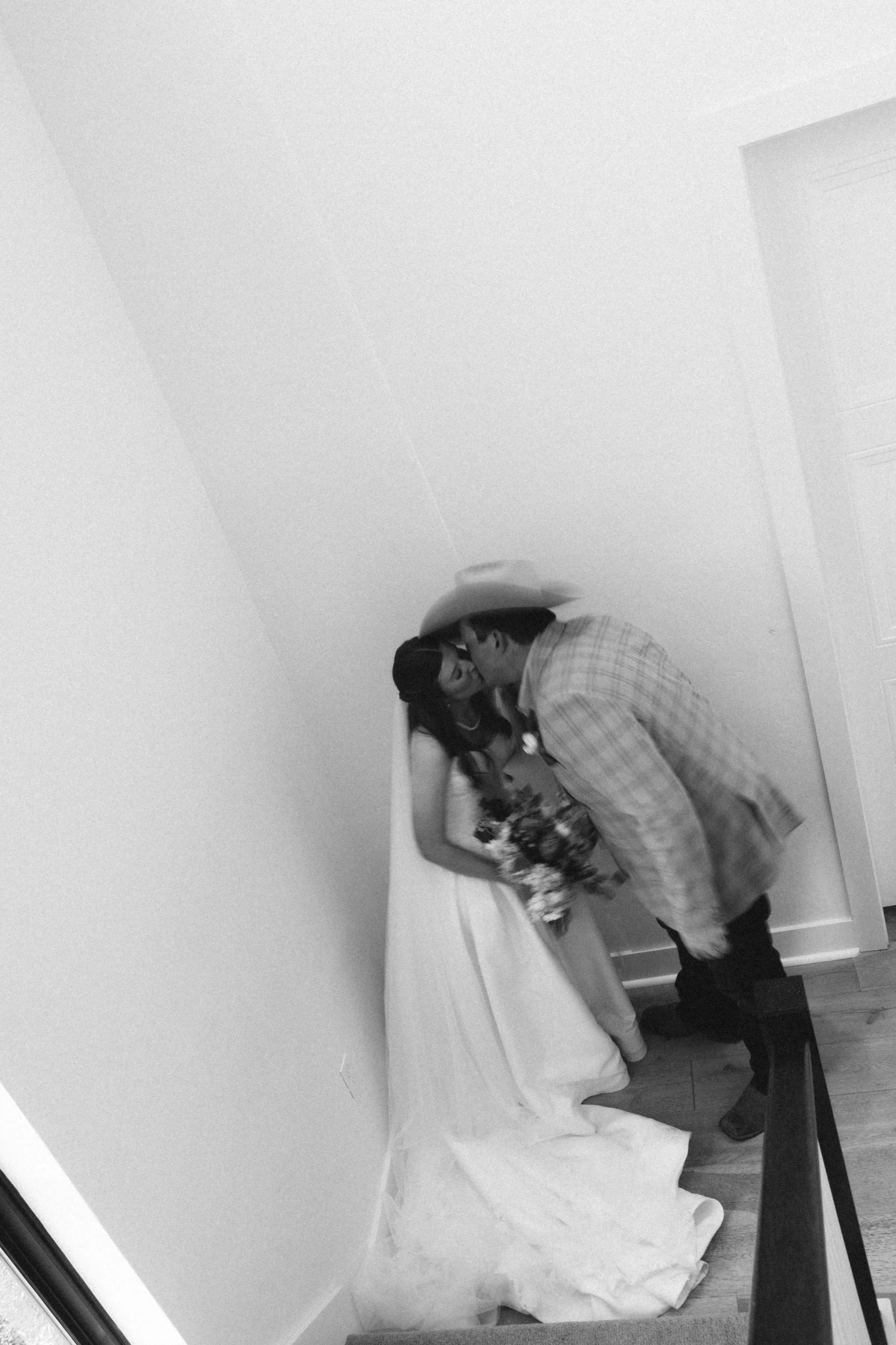 A man wearing a cowboy hat embraces a woman in a floral dress at a table with a white tablecloth in a room with plain walls.