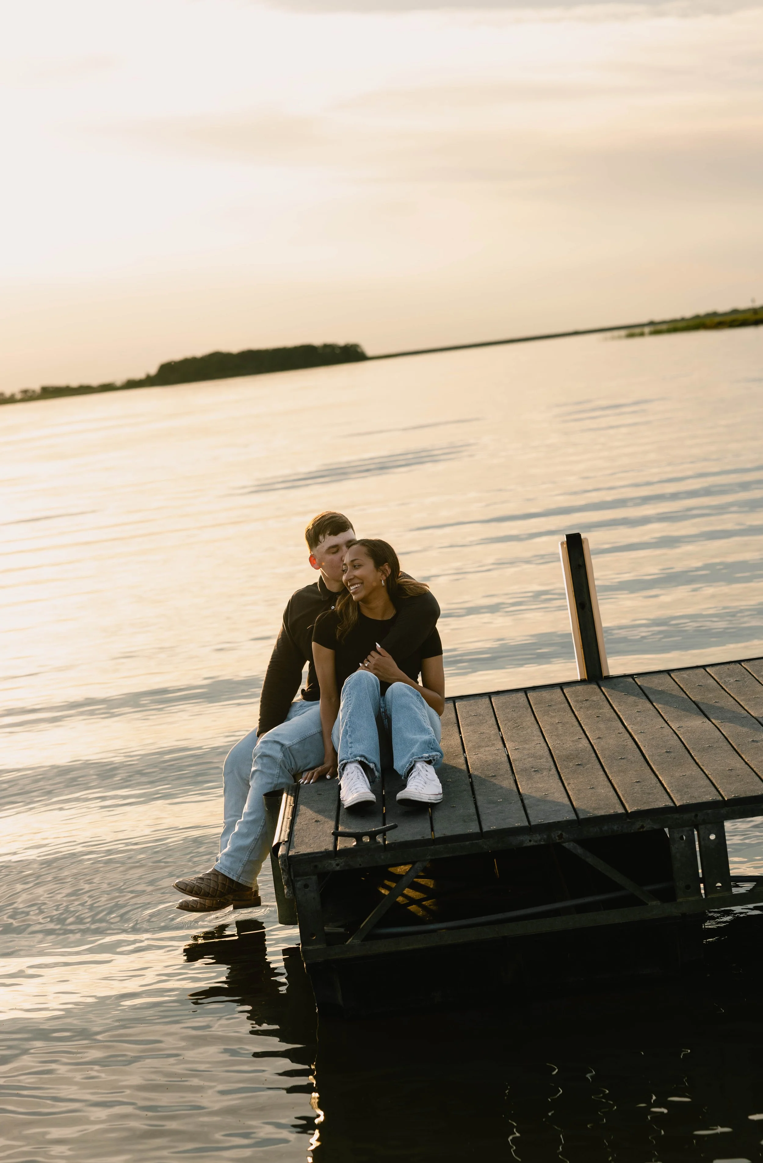 A couple sitting on a dock next to a body of water at sunset, smiling and embracing each other.