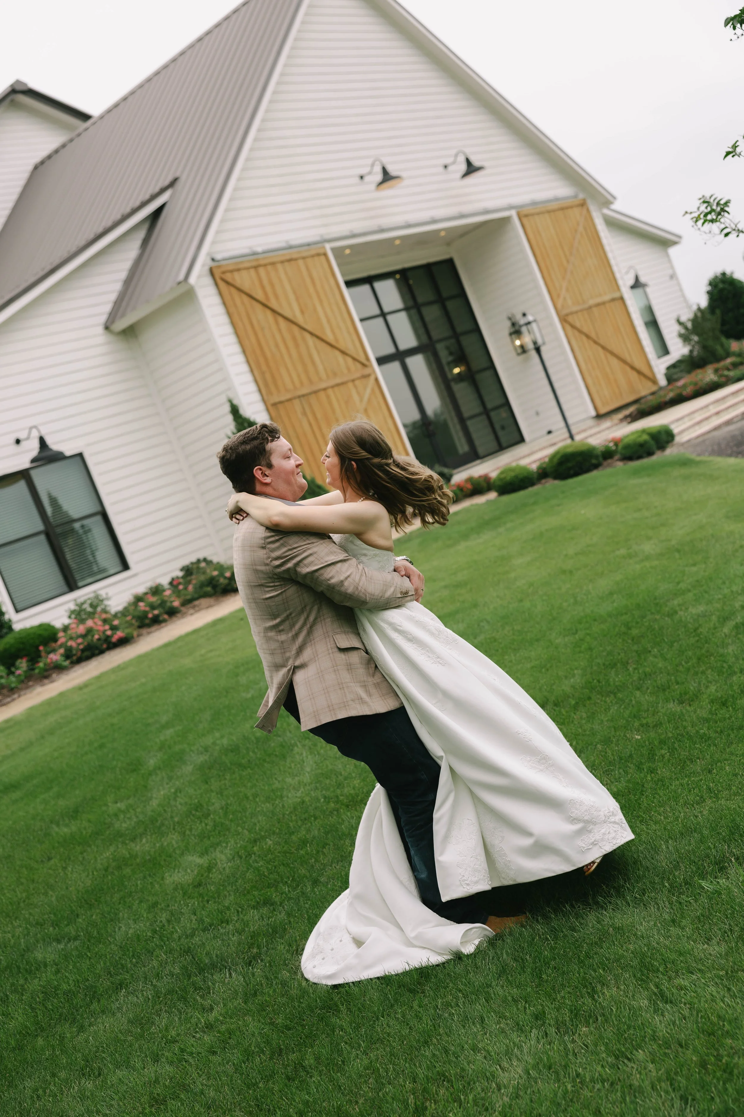 A bride and groom share a dance outside a white house with wooden barn doors on a well-manicured lawn.