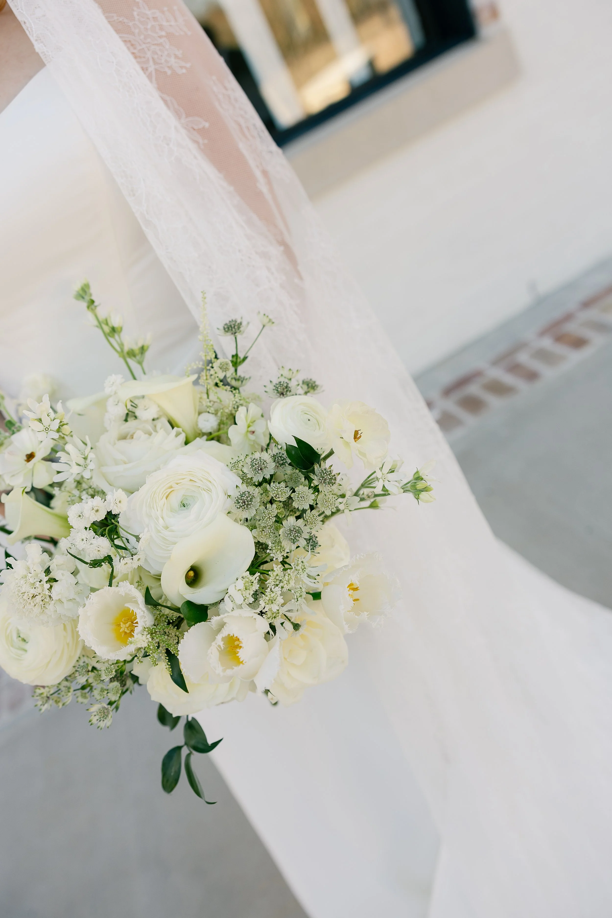 White wedding bouquet with roses, calla lilies, and greenery, placed on a white cloth with a lace veil nearby, against a blurred background.