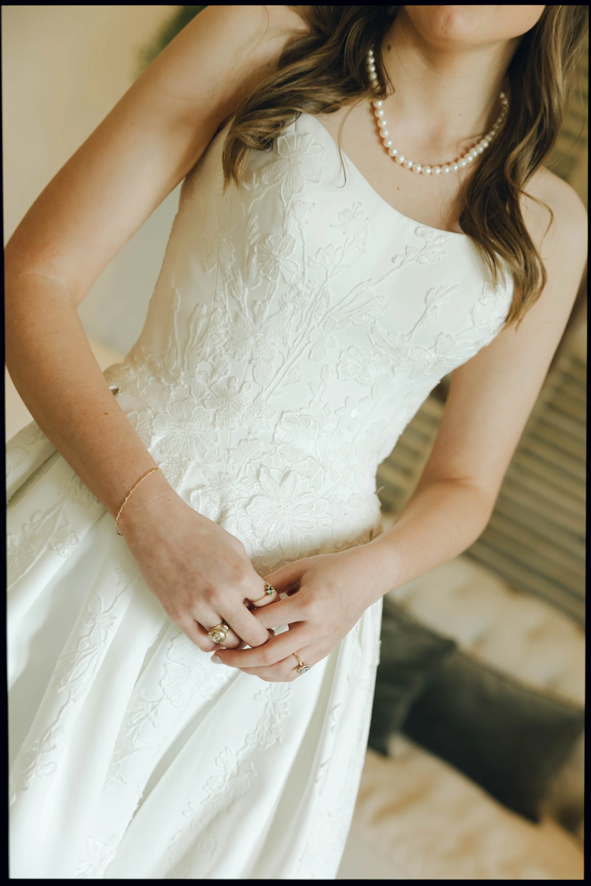 Close-up of a woman in a white wedding dress with floral embroidery, wearing a pearl necklace and multiple rings, standing with her hands clasped in front of her.