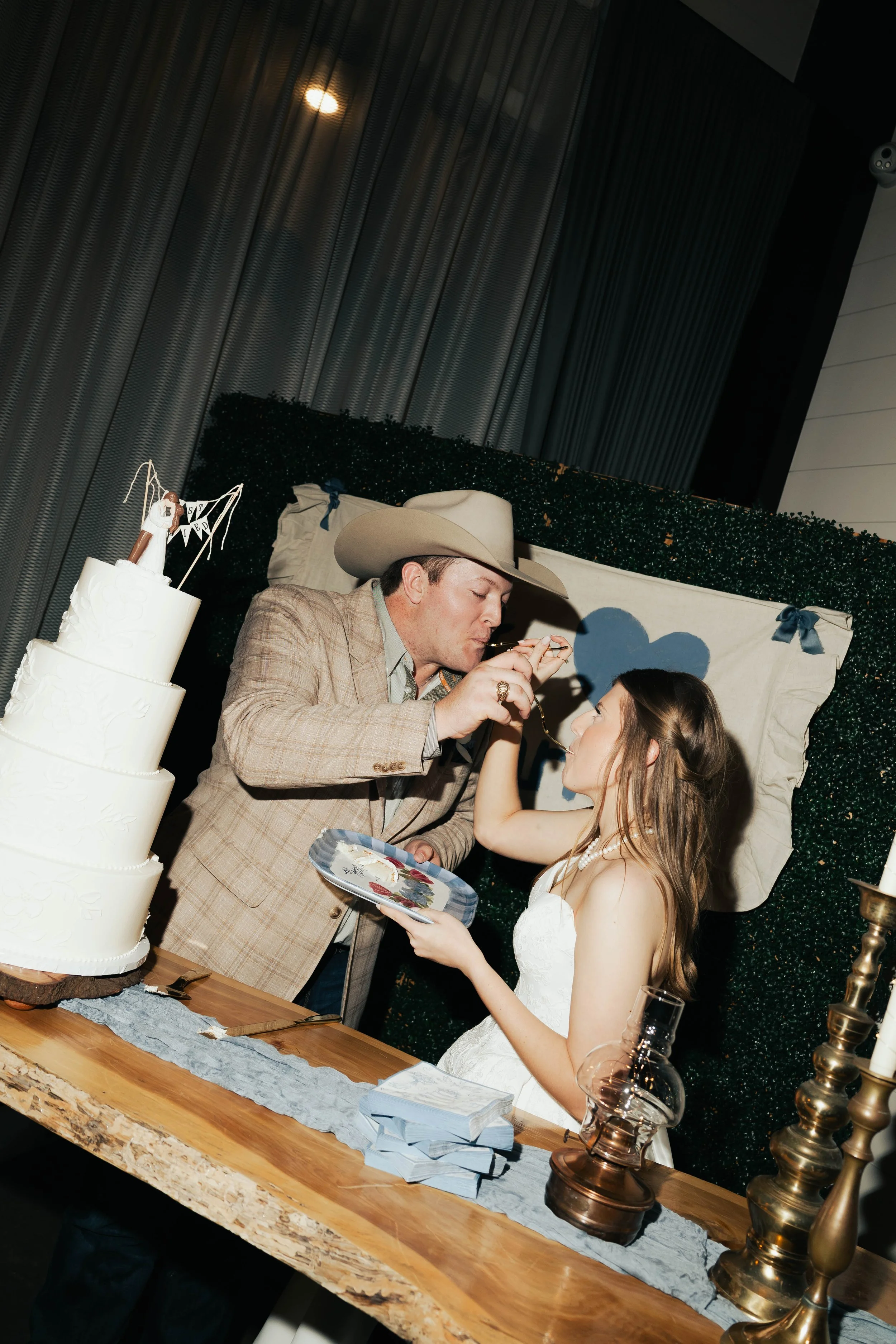 A groom and bride sharing a moment during wedding cake celebration, with a multi-tiered wedding cake nearby, surrounded by decor and candles.