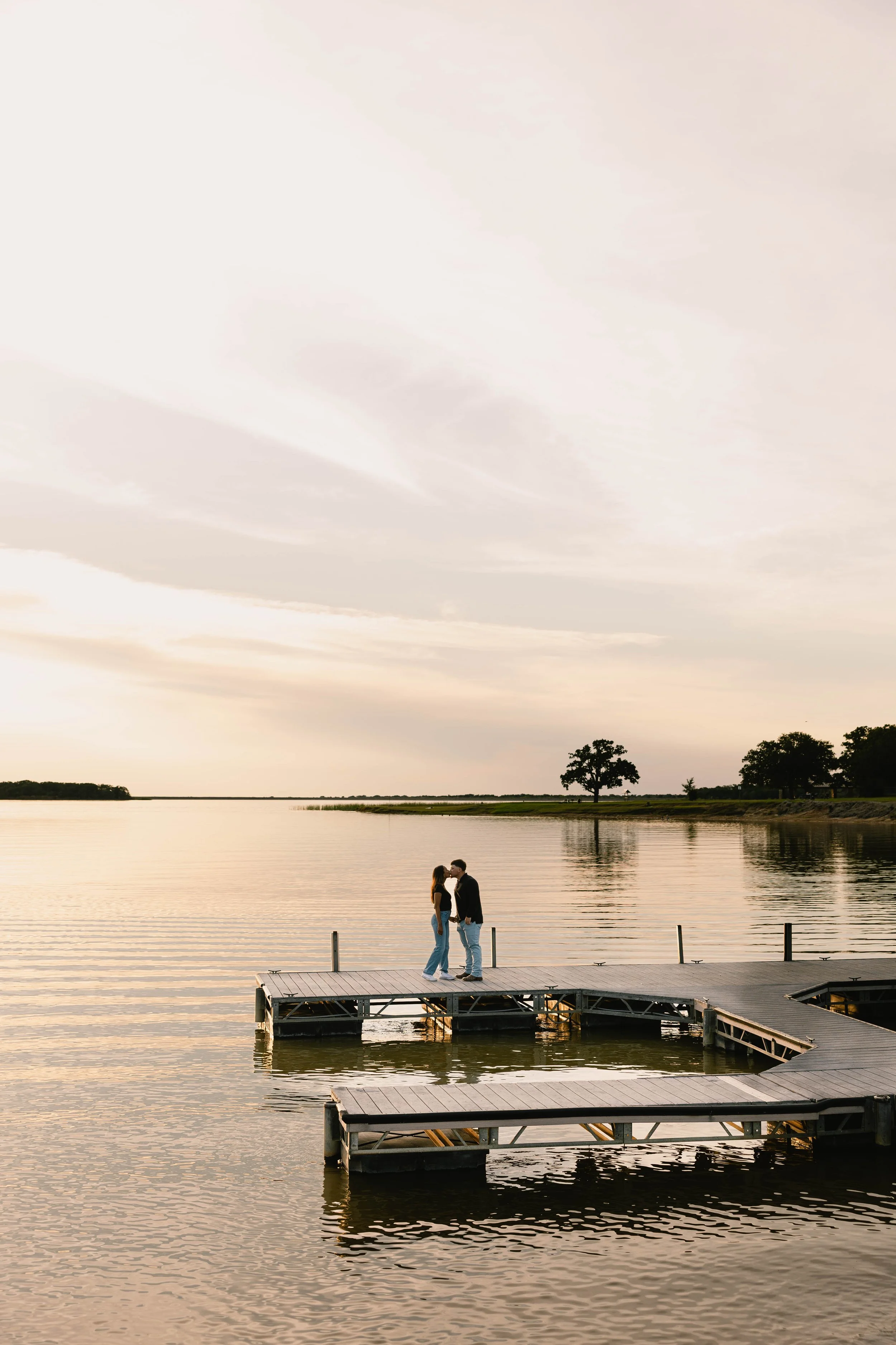 A couple is standing on a dock by a lake, about to kiss, during sunset with calm water and trees in the background.