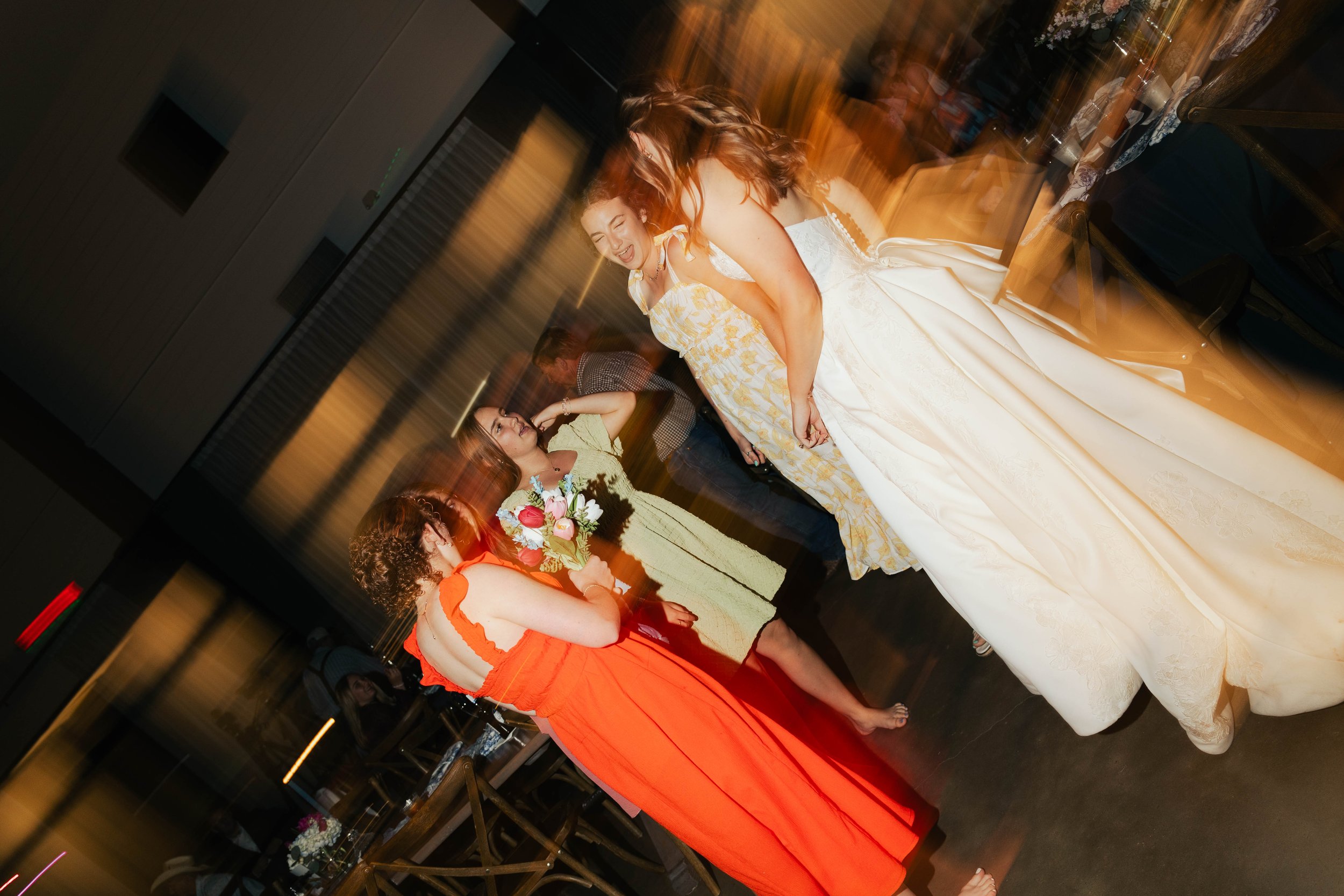 Two women are dancing and smiling at a party, with one wearing a white dress and the other wearing an orange dress. Other guests can be seen in the background, and the setting appears to be a decorated indoor venue.