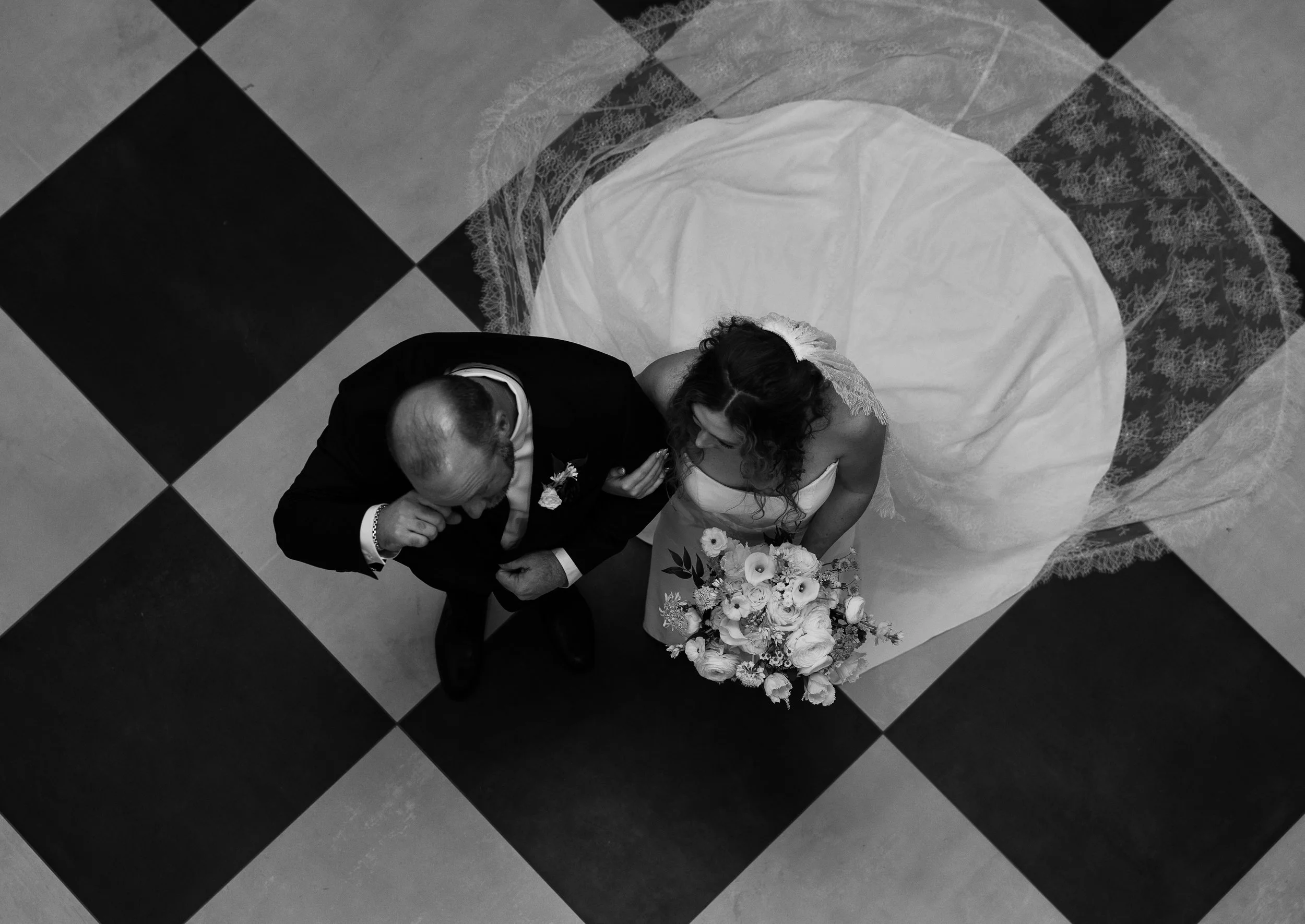 Black and white aerial view of a bride and groom on a checkered floor, with the bride holding a bouquet of flowers.