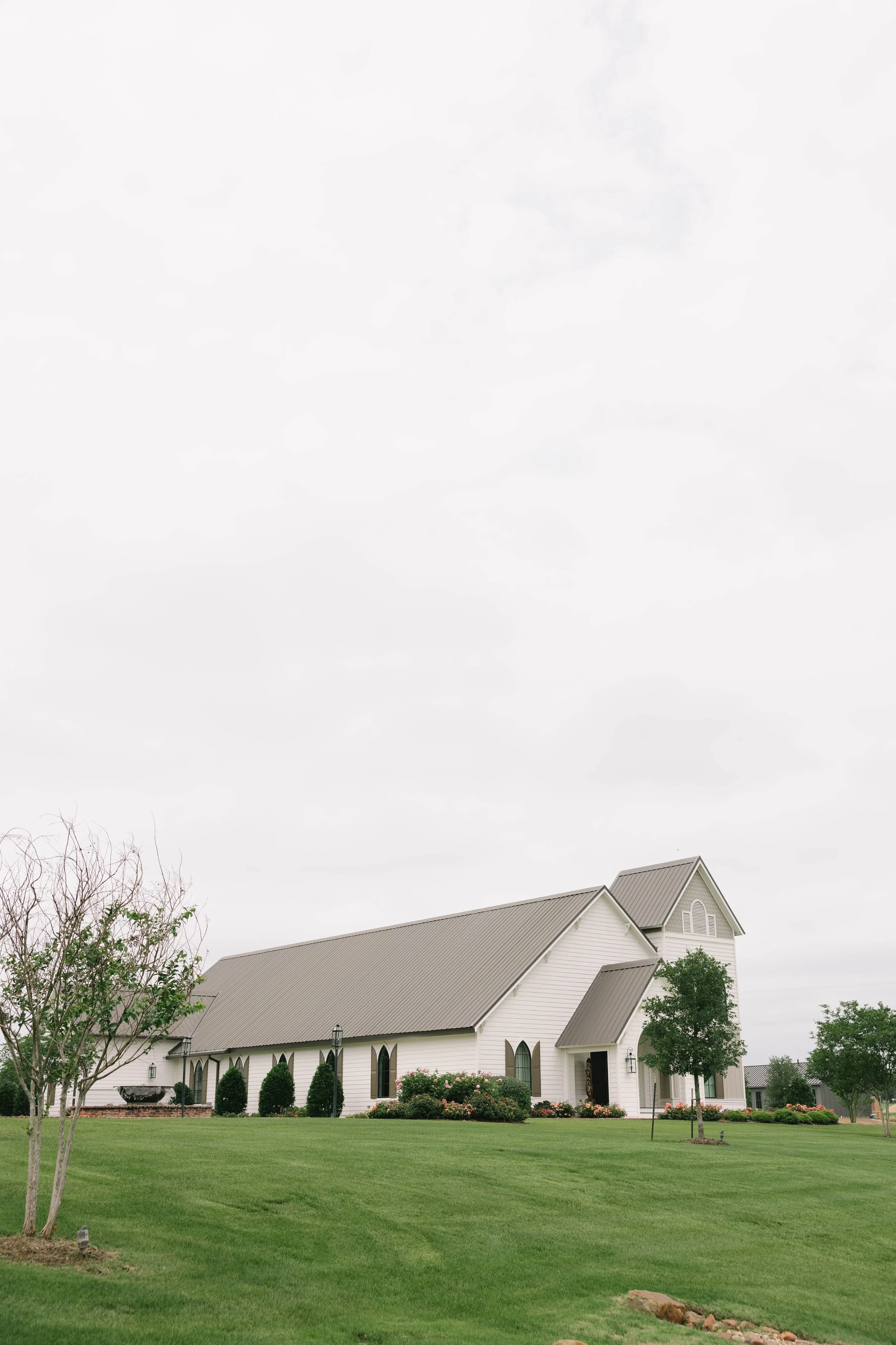 A white church with a tall steeple and arched windows, surrounded by a green lawn, trees, and shrubs, under a cloudy sky.