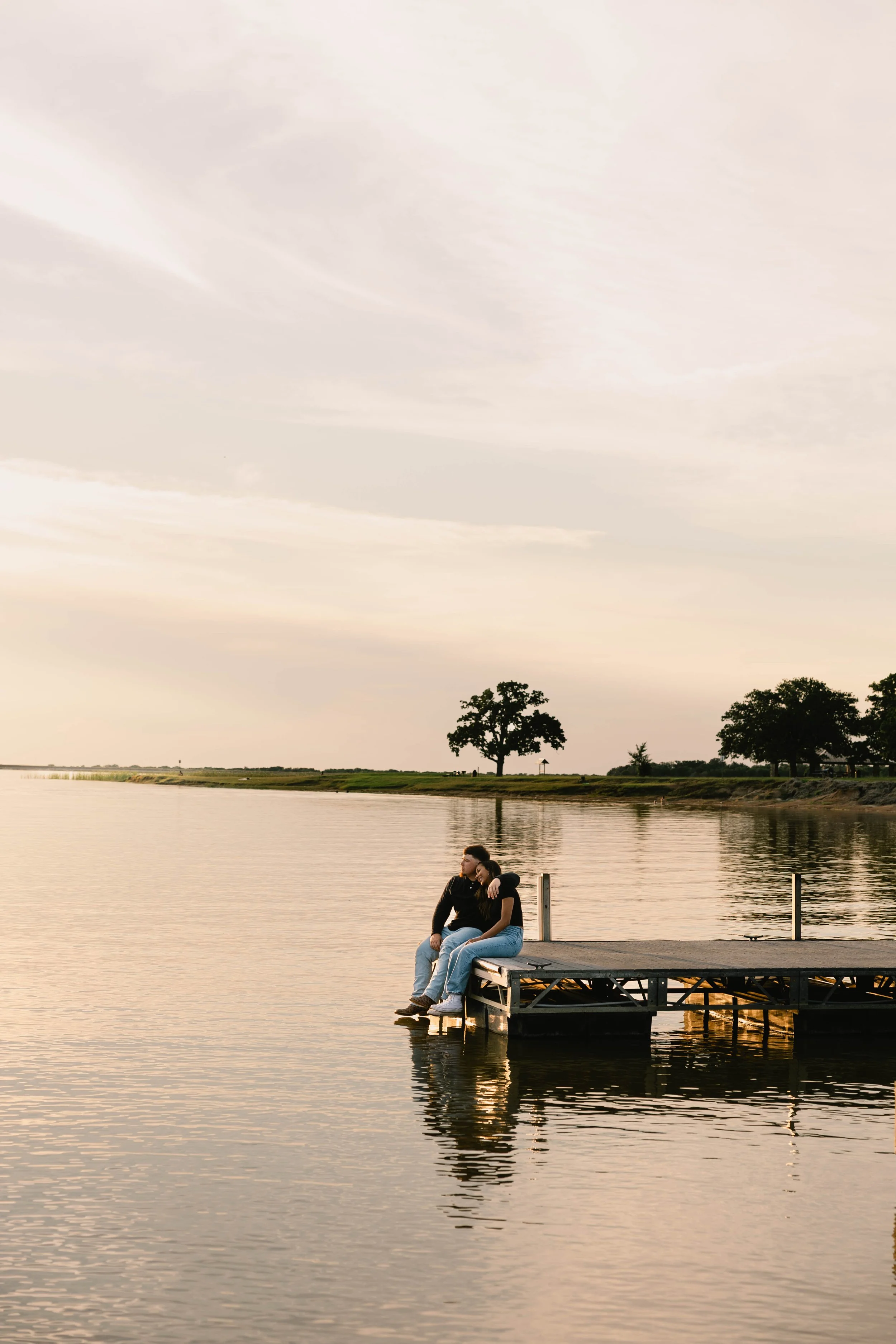 A couple sitting on a dock by a lake at sunset, with trees in the background and calm rippling water.