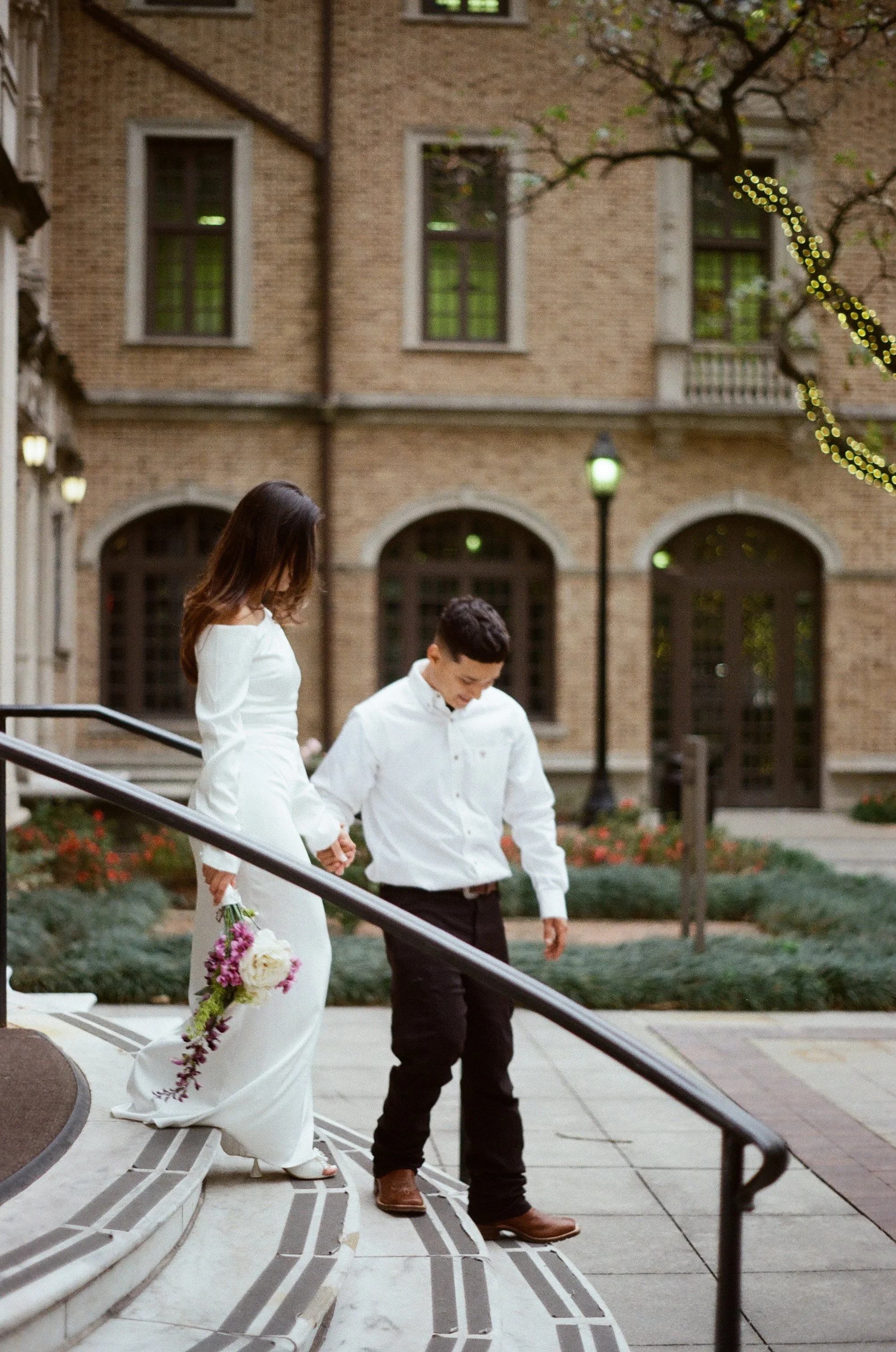 A bride and groom walking down the stairs outside a brick building, holding hands, the bride holding a bouquet of flowers, and dressed in a white gown, the groom in a white shirt and black pants.
