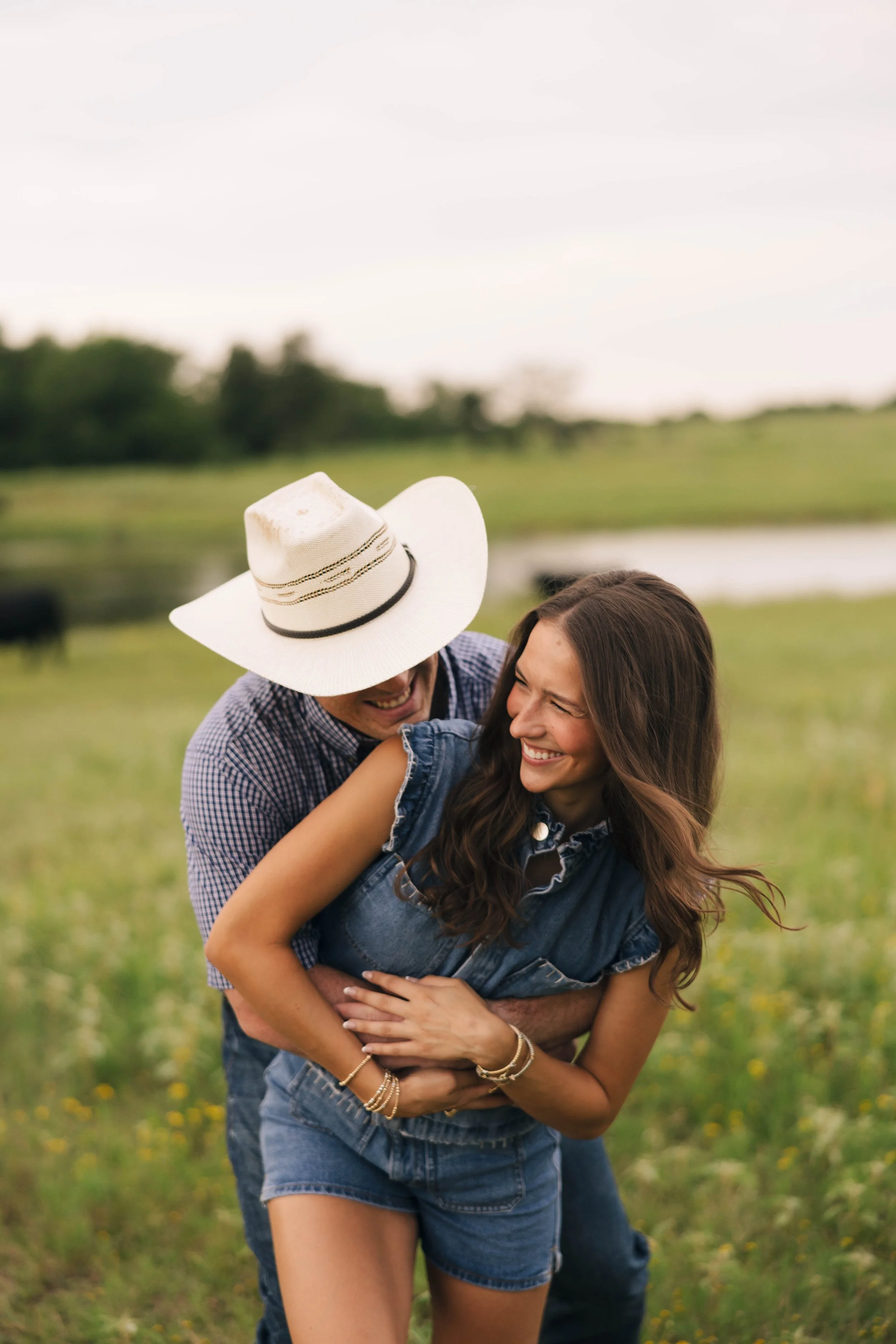 A man in a plaid shirt and a white straw hat playfully hugging a woman in a denim shirt and shorts in a grassy field, both laughing and enjoying each other's company.