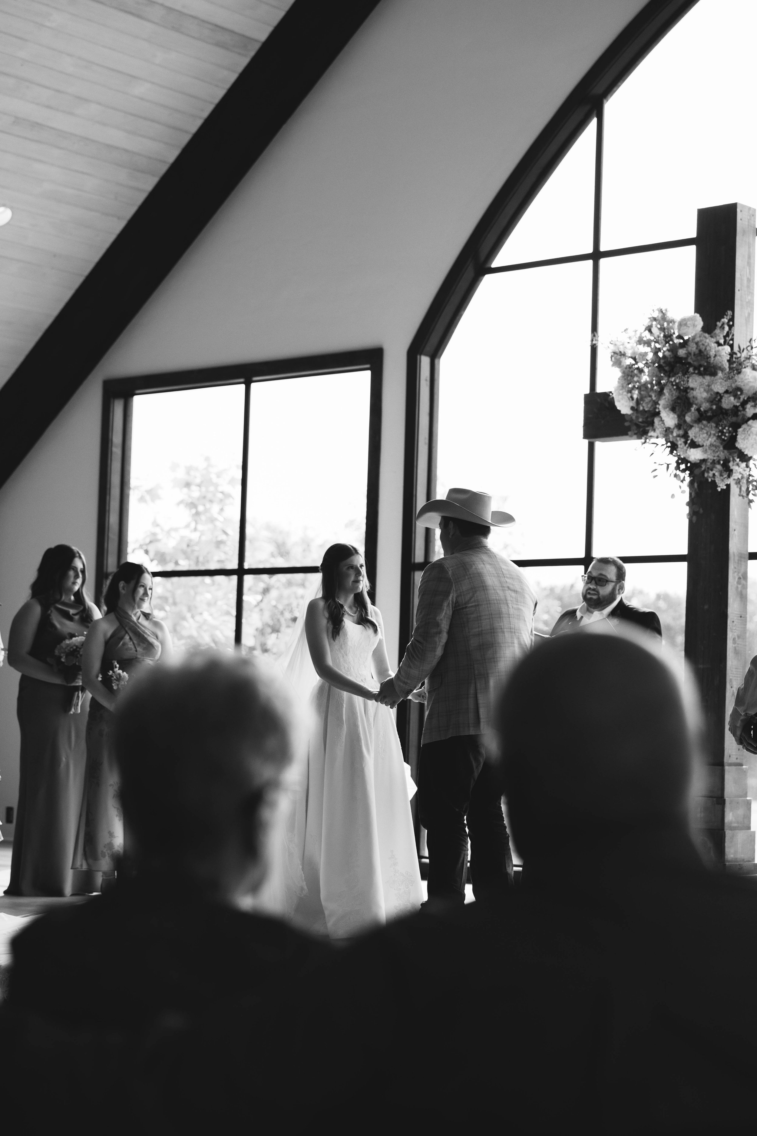 A black and white photo of a wedding ceremony inside a church with large windows and a cross decorated with flowers in the background. The bride and groom are holding hands, facing each other, with bridesmaids and groomsmen on either side.
