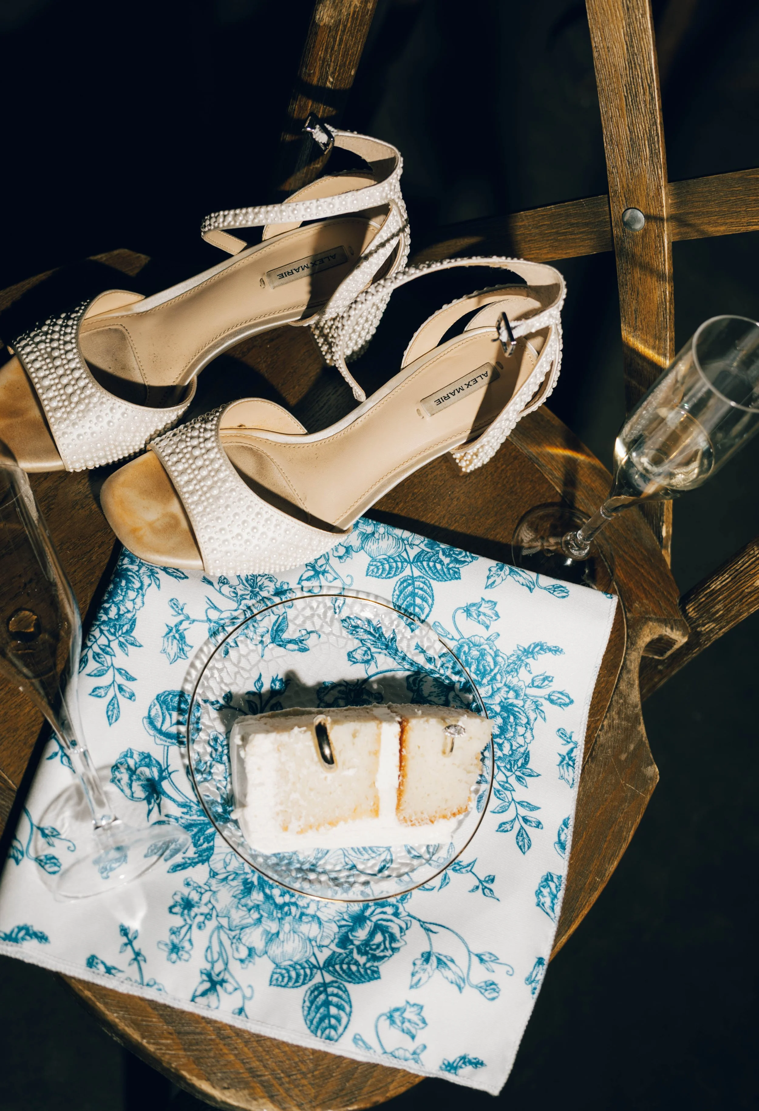 A pair of white high-heeled shoes with pearl embellishments resting on a wooden chair, a slice of cake with white frosting and a caramel layer on a blue and white floral patterned napkin, and two empty champagne glasses on a dessert plate.