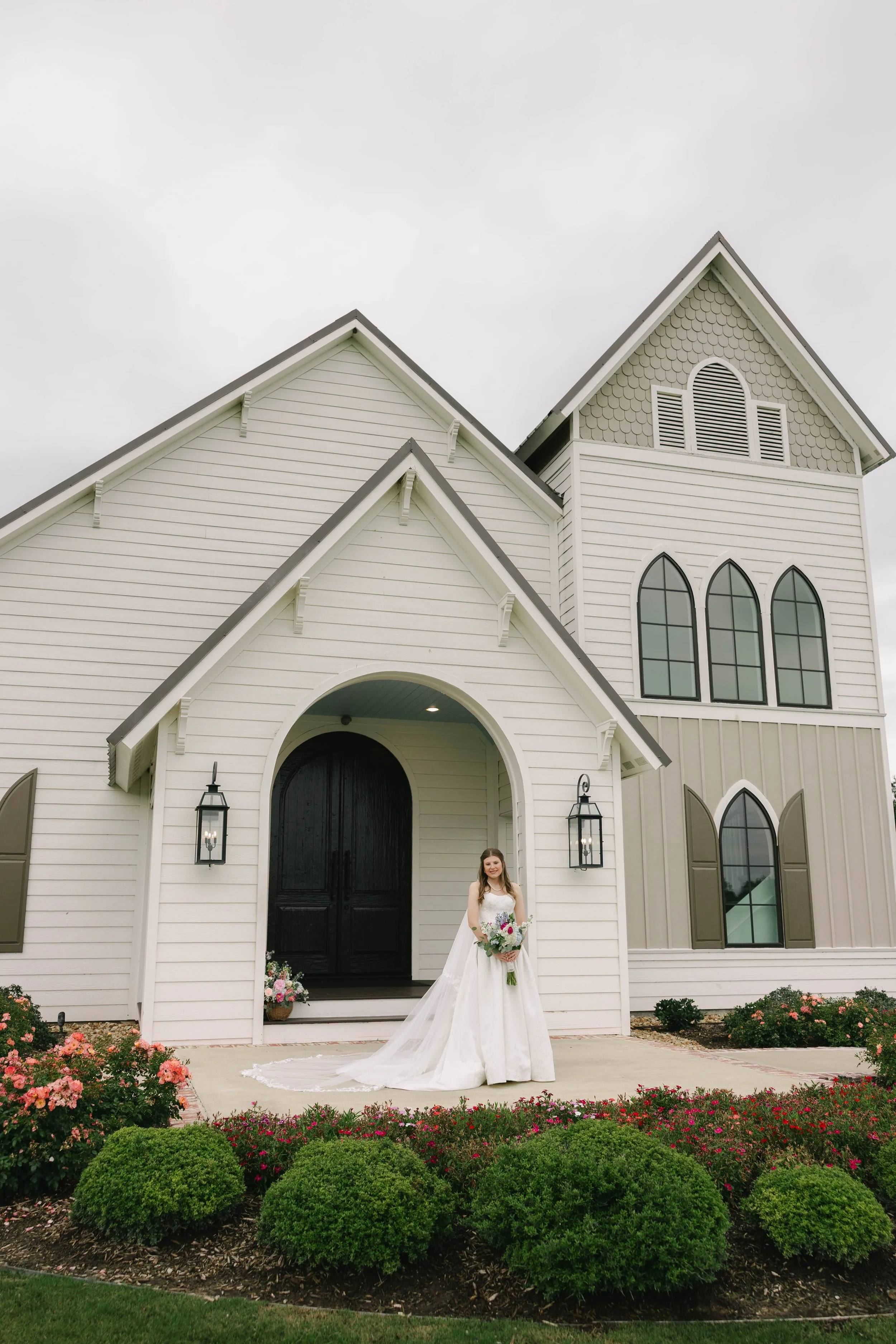 A bride standing in front of a white church holding a bouquet of flowers. The church has black doors, arched windows, and flower beds with pink flowers and green bushes.