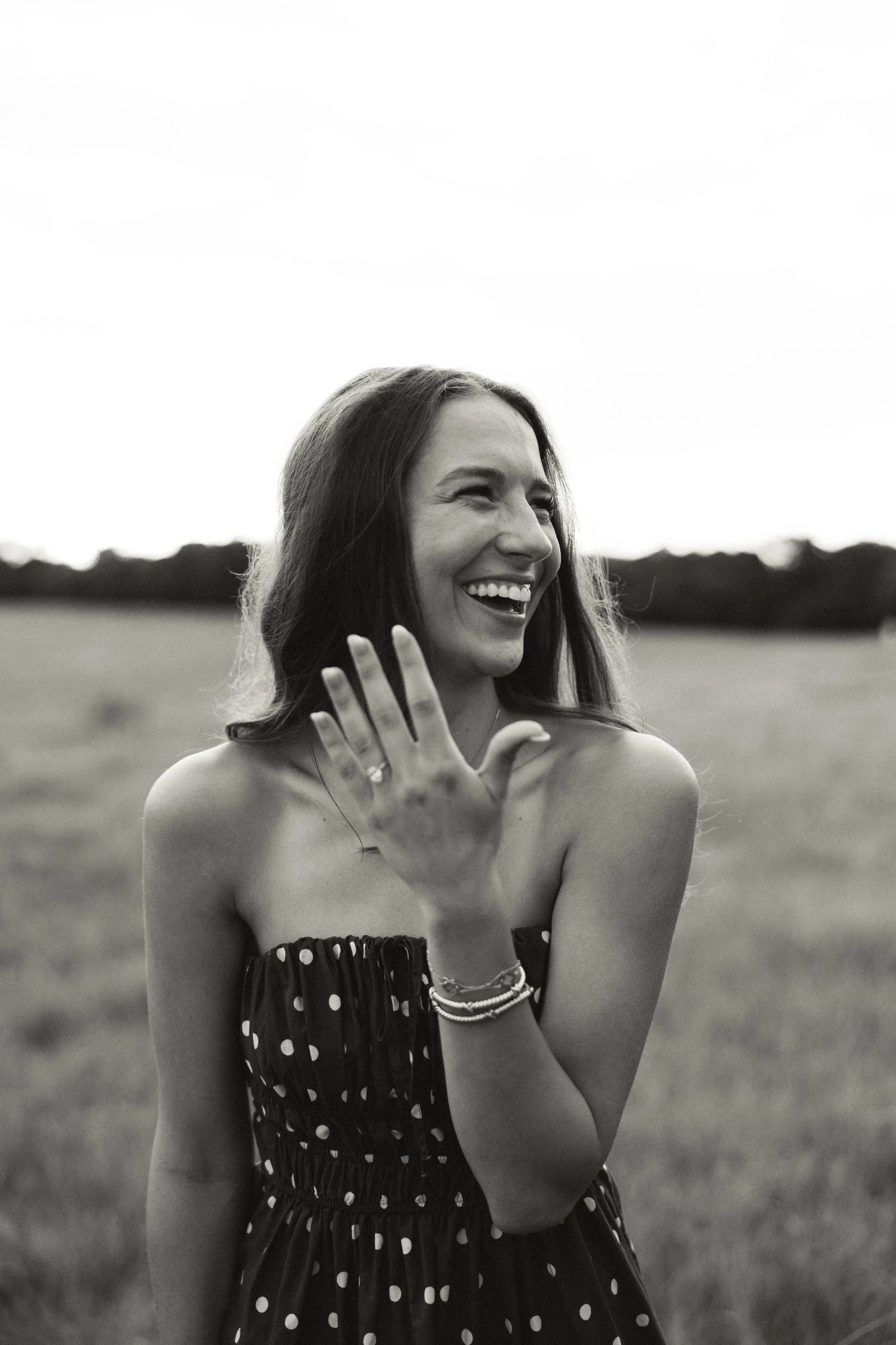 A woman laughing outdoors in a field, wearing a strapless polka dot dress, with her hand raised showing a ring on her finger.