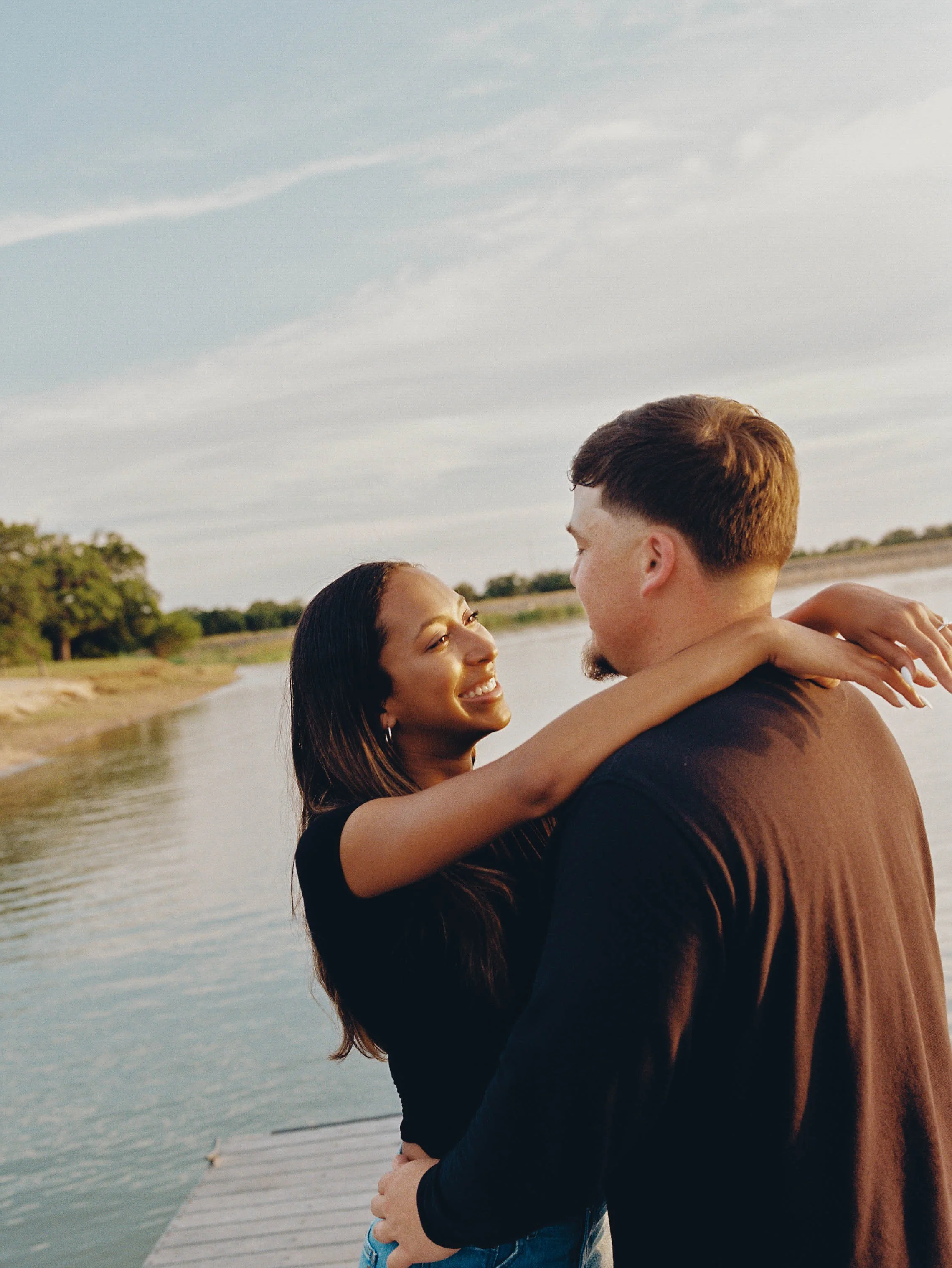 A couple stands close together outdoors near a body of water, smiling and looking into each other's eyes.