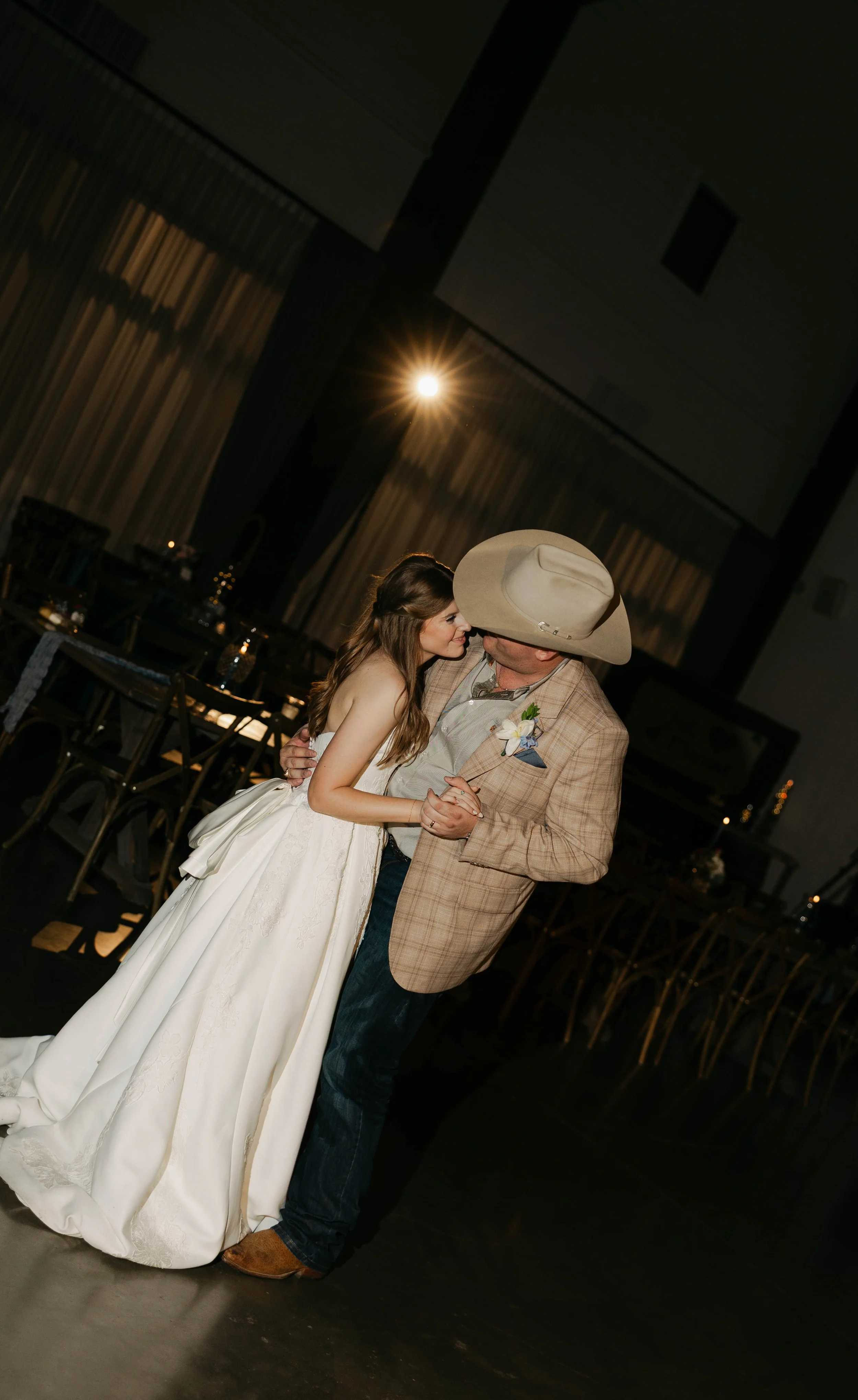 A woman in a wedding dress dancing with a man wearing a cowboy hat and plaid jacket in a dimly lit room decorated for a wedding reception.