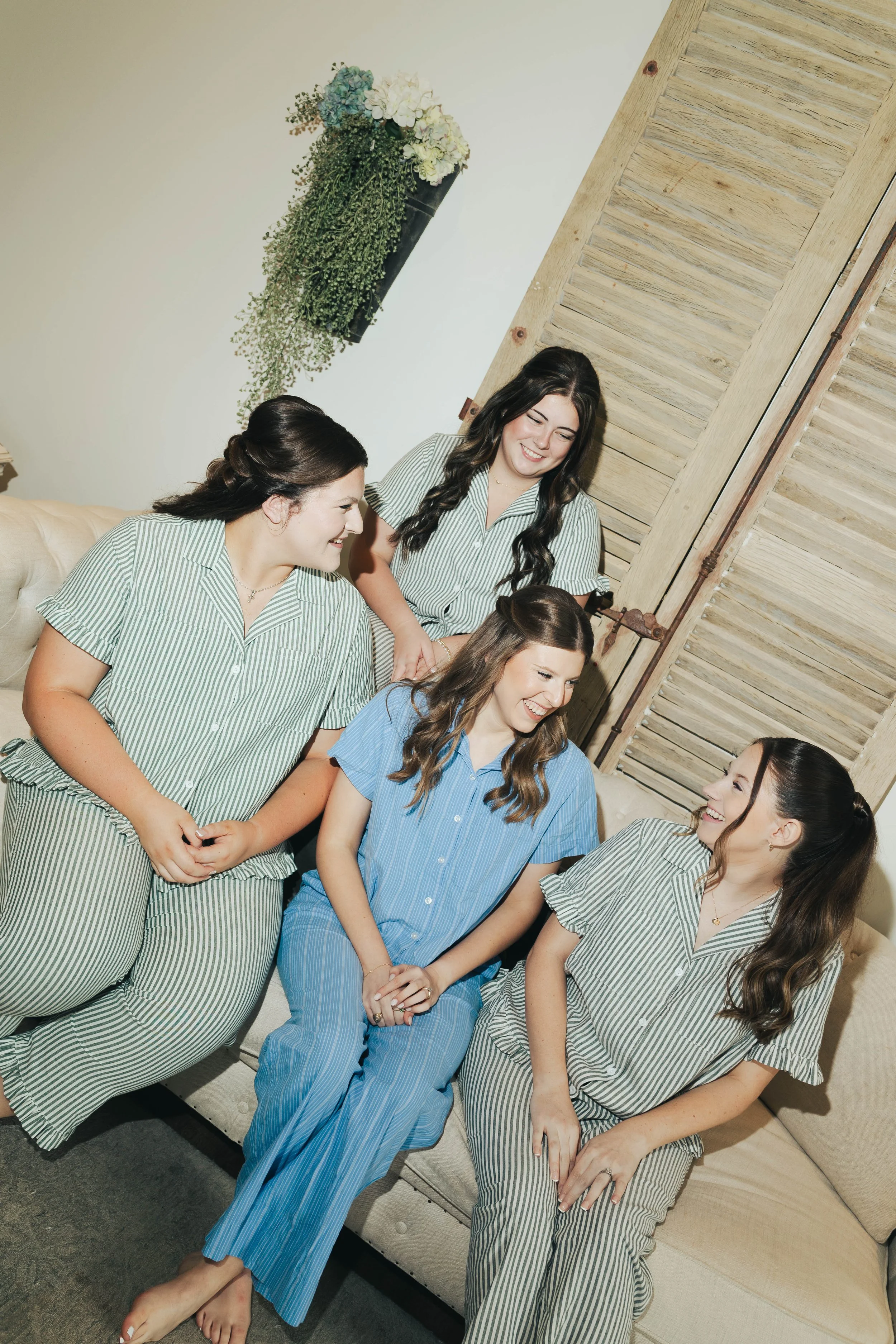 Group of six women sitting and standing close together, smiling and talking, wearing matching striped pajamas in green, blue, and beige tones, in a cozy room with wooden wall paneling and a floral wall hanging.