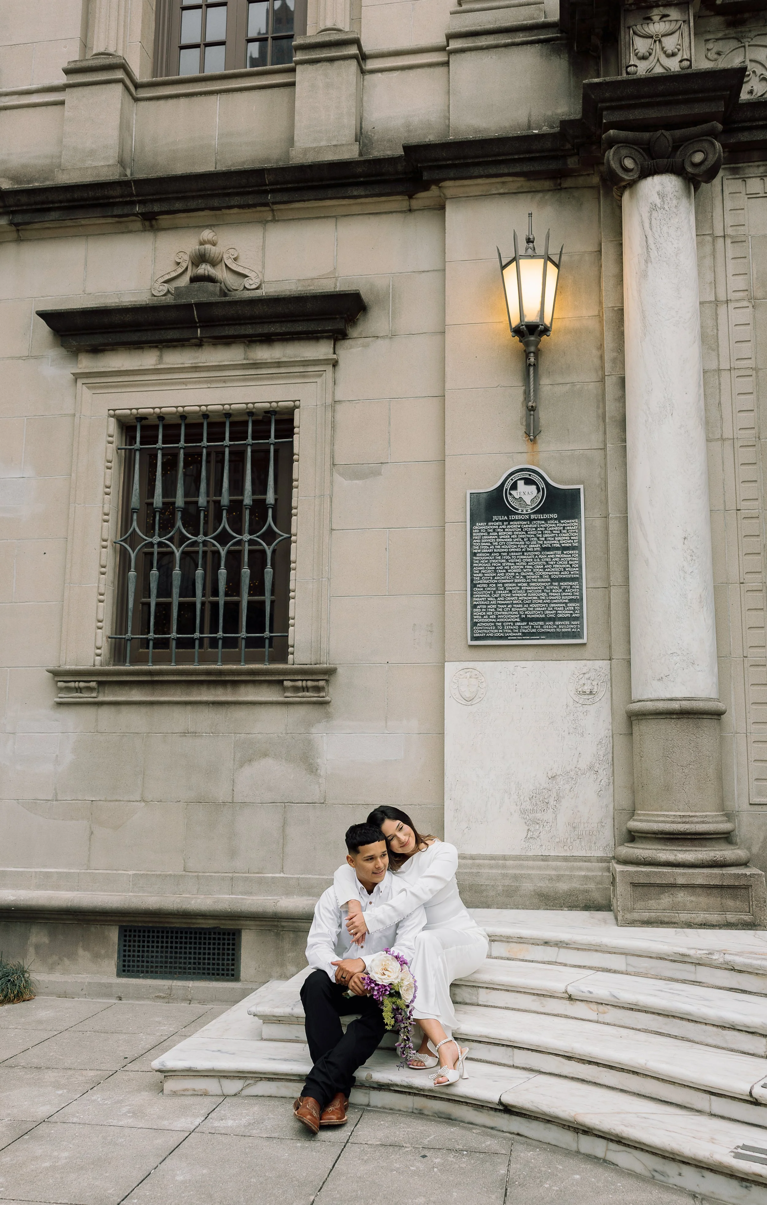 Couple dressed in white sitting on marble steps outside a historic building with a lamp, a window with iron bars, and a plaque on the wall.