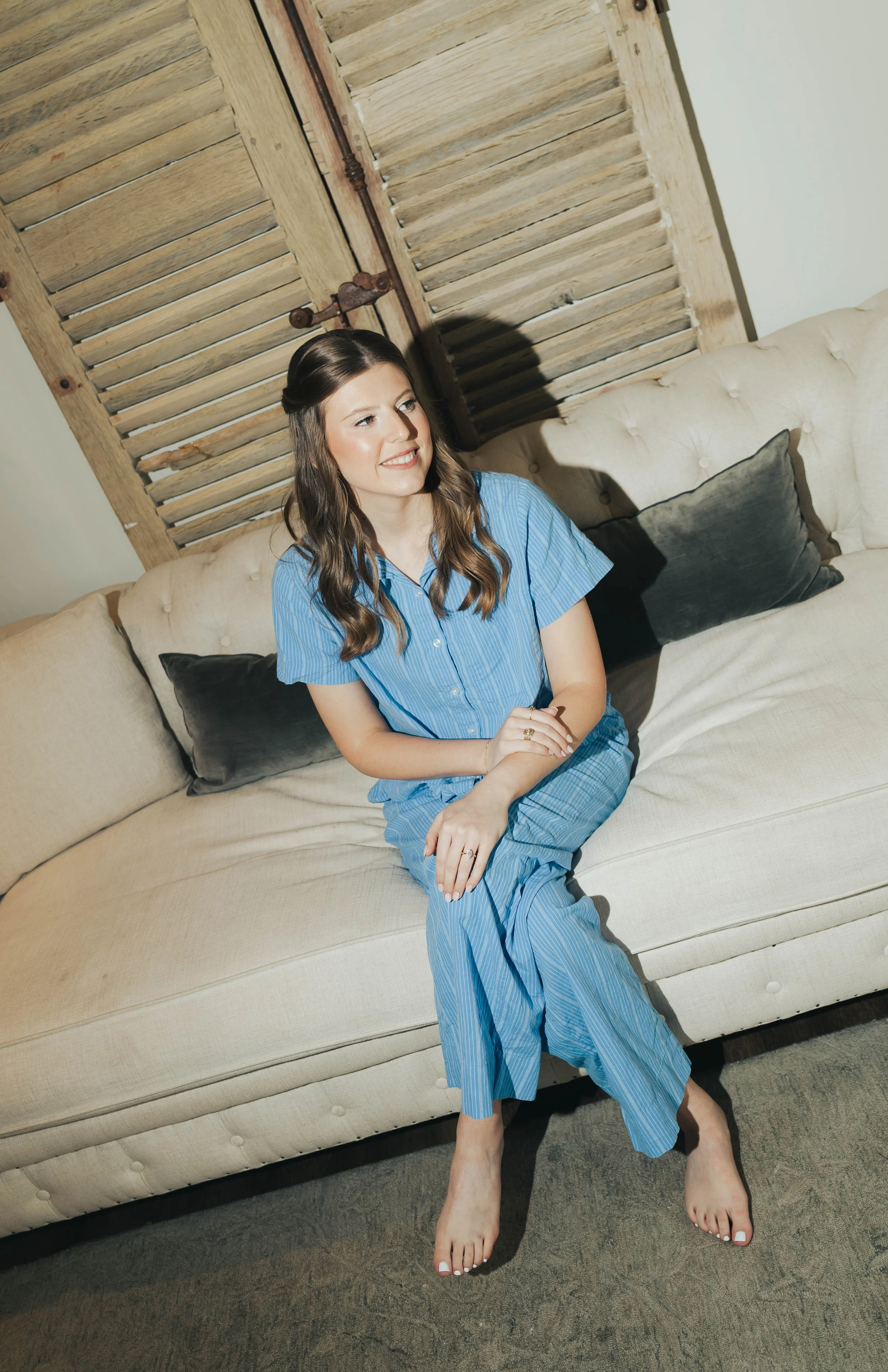 A woman with long brown hair, wearing blue pajamas, sitting on a beige couch with her legs and feet visible, in a room with a wooden louvered door and a white wall.