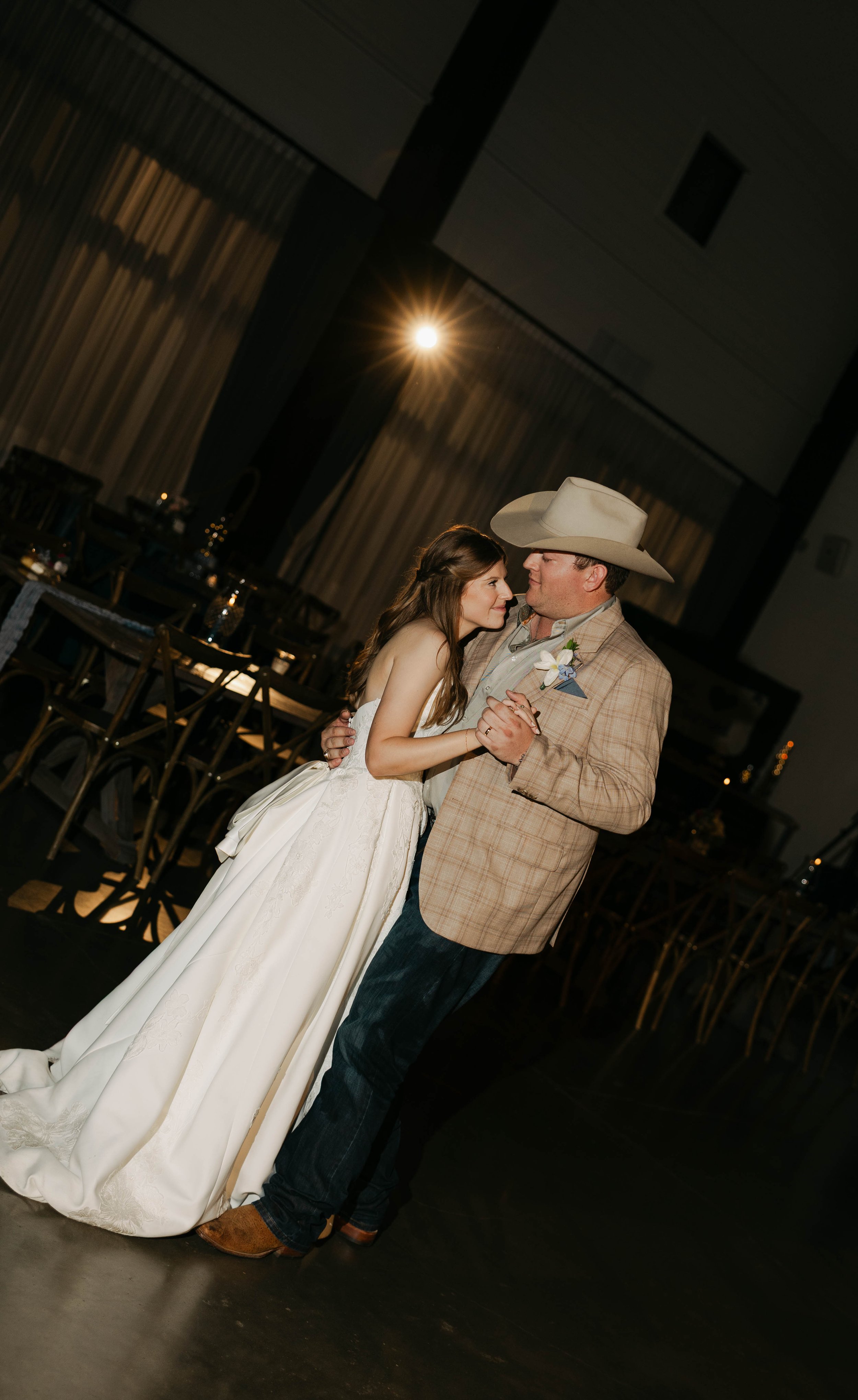 A bride in a white wedding dress dancing with an older man wearing a cowboy hat and a plaid blazer in a decorated reception hall.
