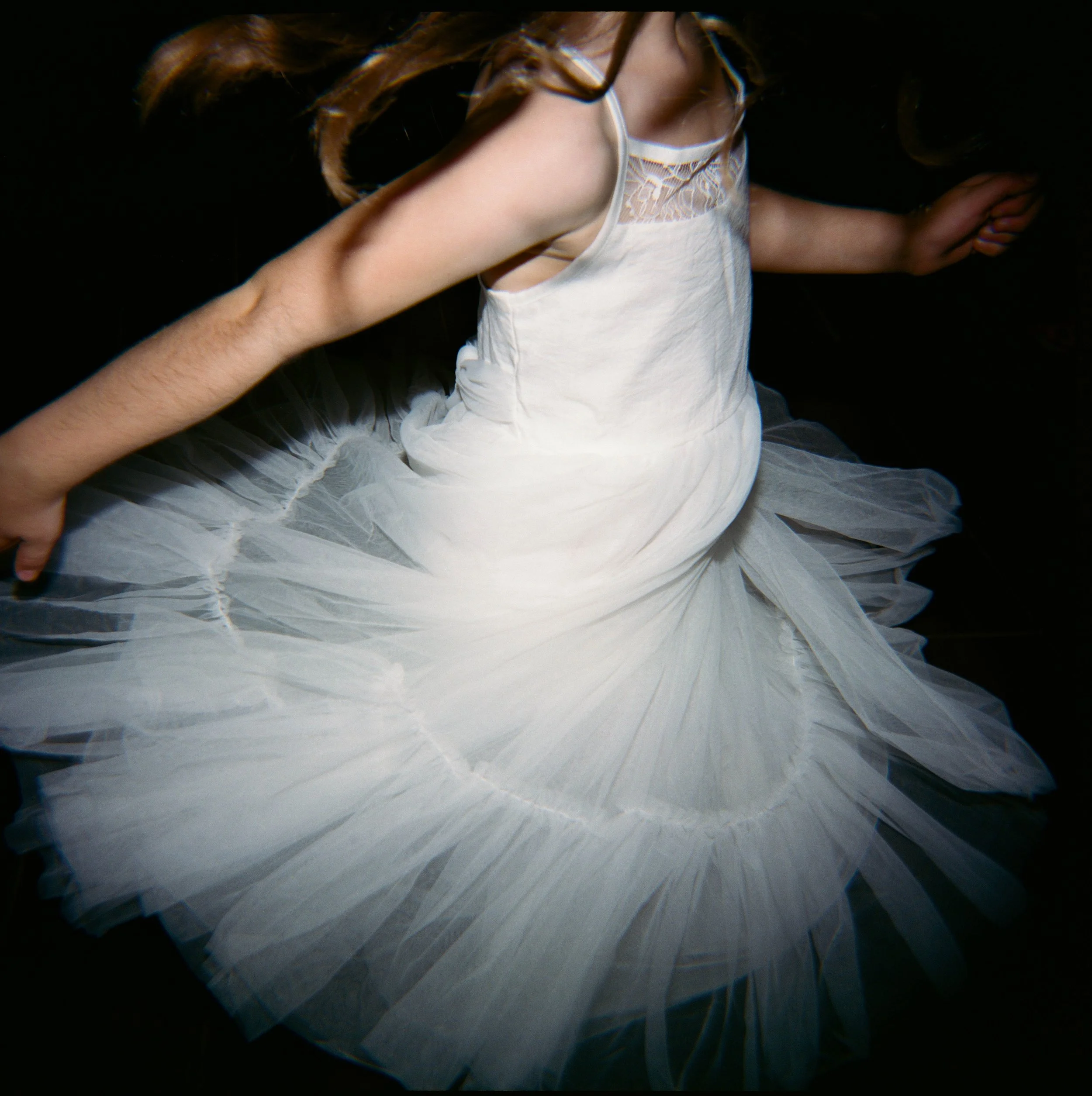 A person in a white dress with a tulle skirt and lace details on the bodice, sitting or kneeling with arms extended to the sides against a dark background.