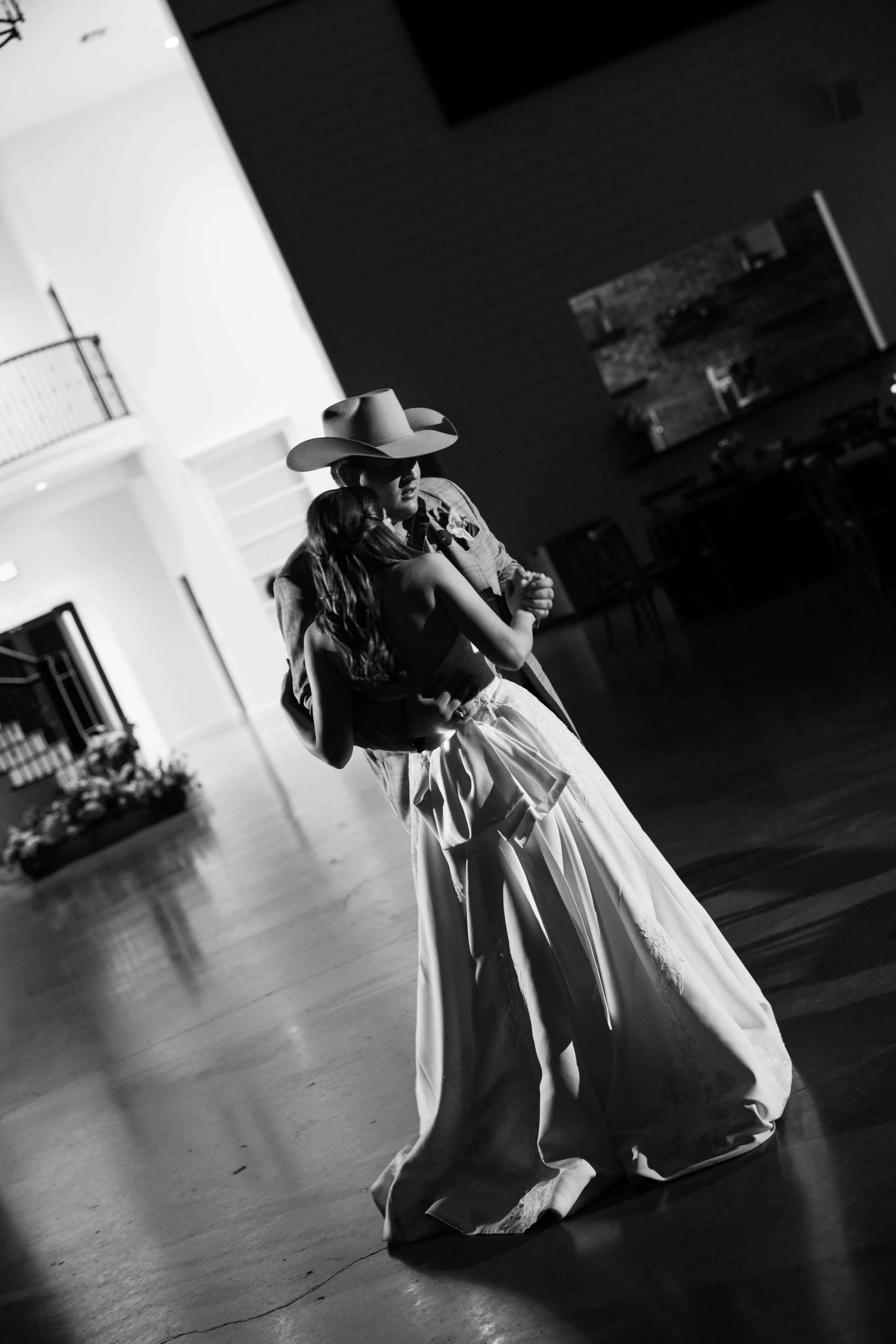 A black and white photo of a bride and groom sharing a dance at their wedding reception. The groom is wearing a cowboy hat. Deep in the Heart Farms is a charming wedding venue in Brenham, Texas, known for its scenic countryside views, elegant event s