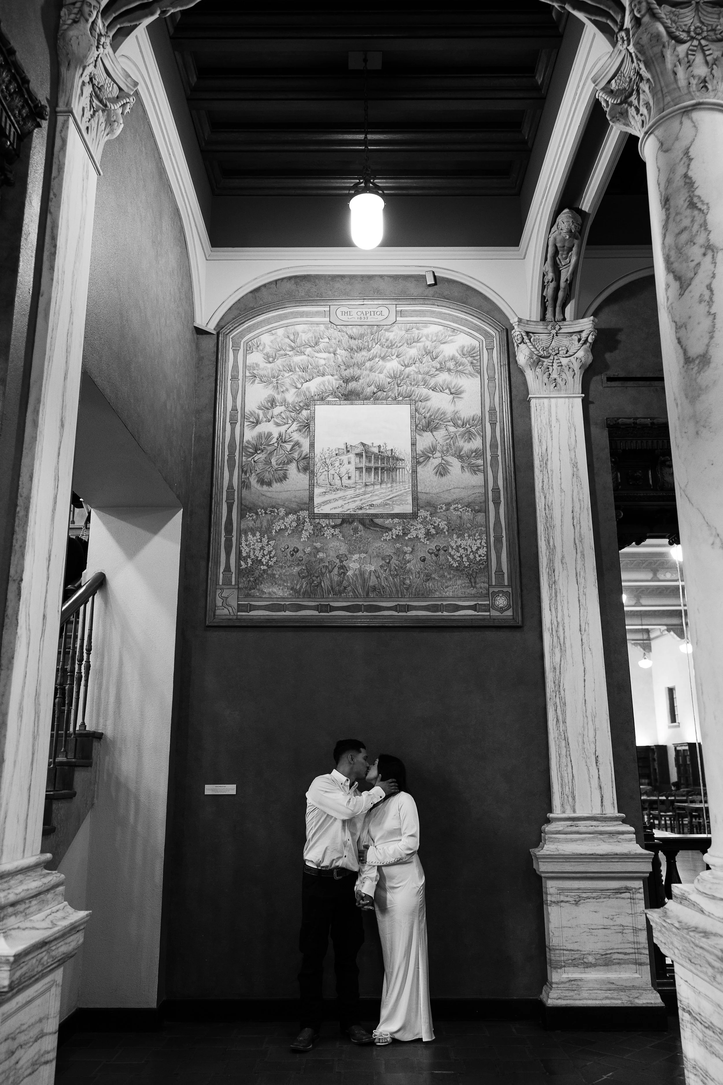 Two people in formal attire embracing and kissing inside a grand hall with ornate columns and artwork on the wall.