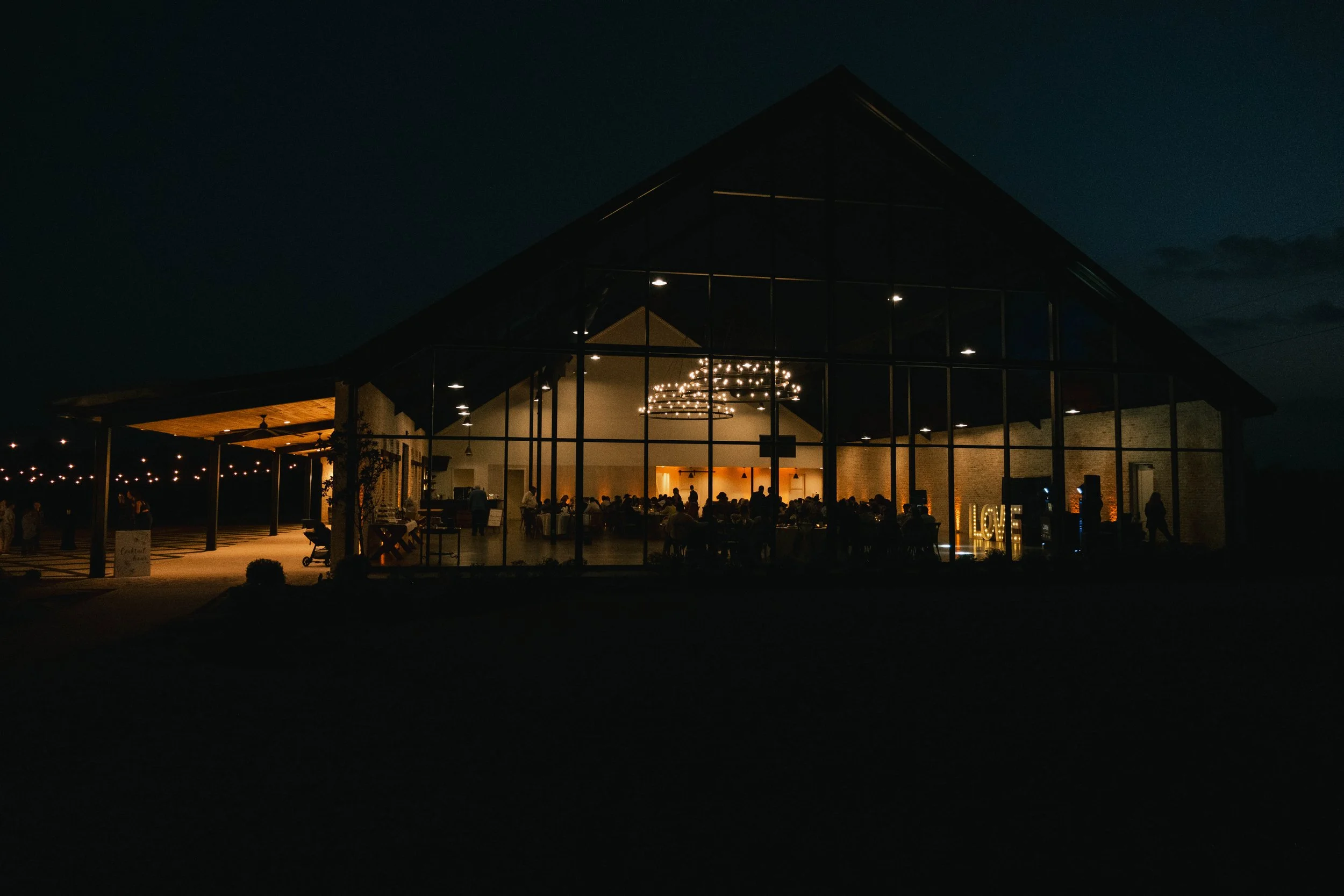 Night scene of a large glass-walled event hall decorated with string lights, with people gathered inside and outside, and a illuminated "LOVE" sign near the entrance.