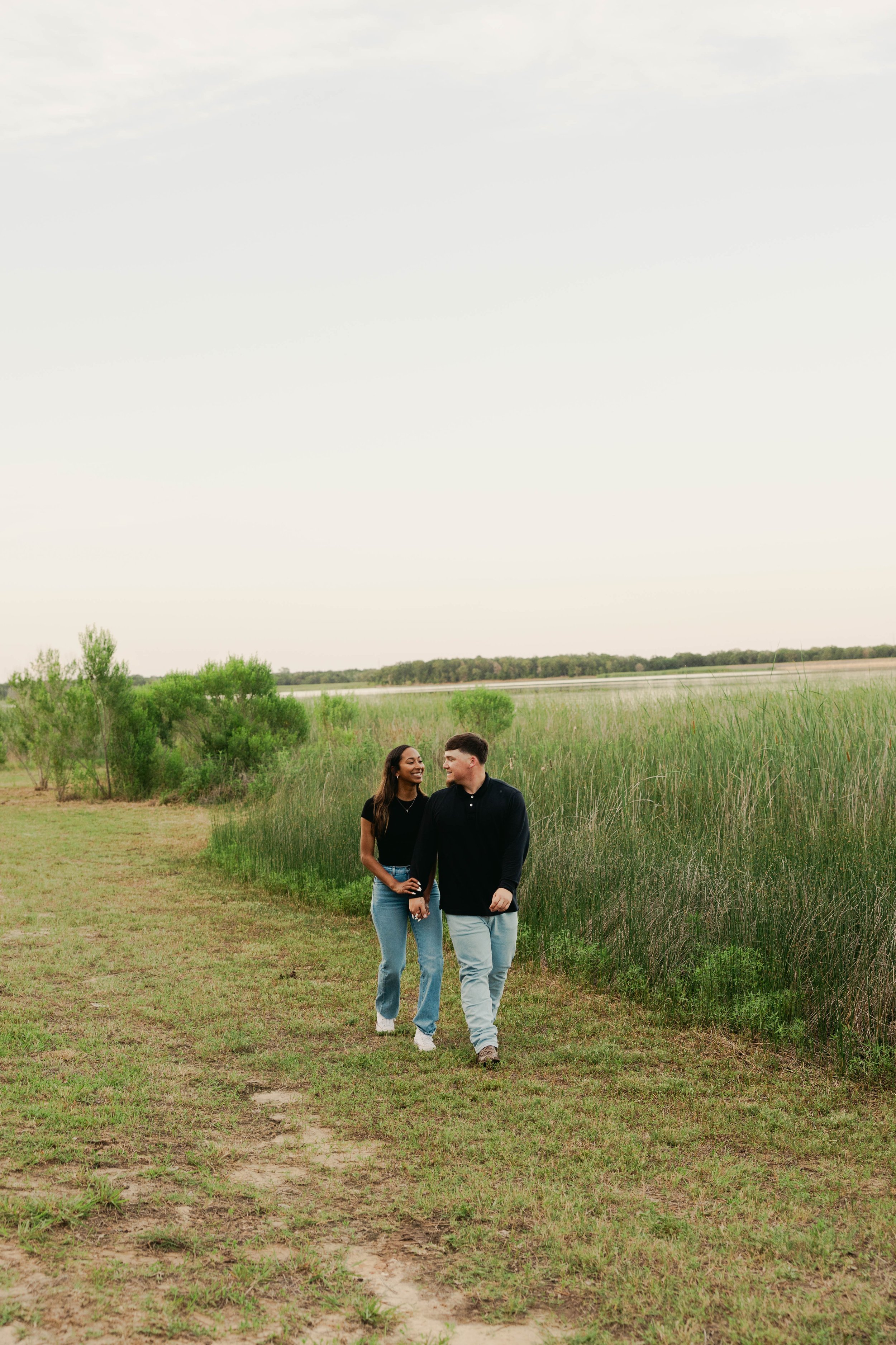 A couple walking along a grassy path in a natural outdoor setting with green shrubs and trees in the background.