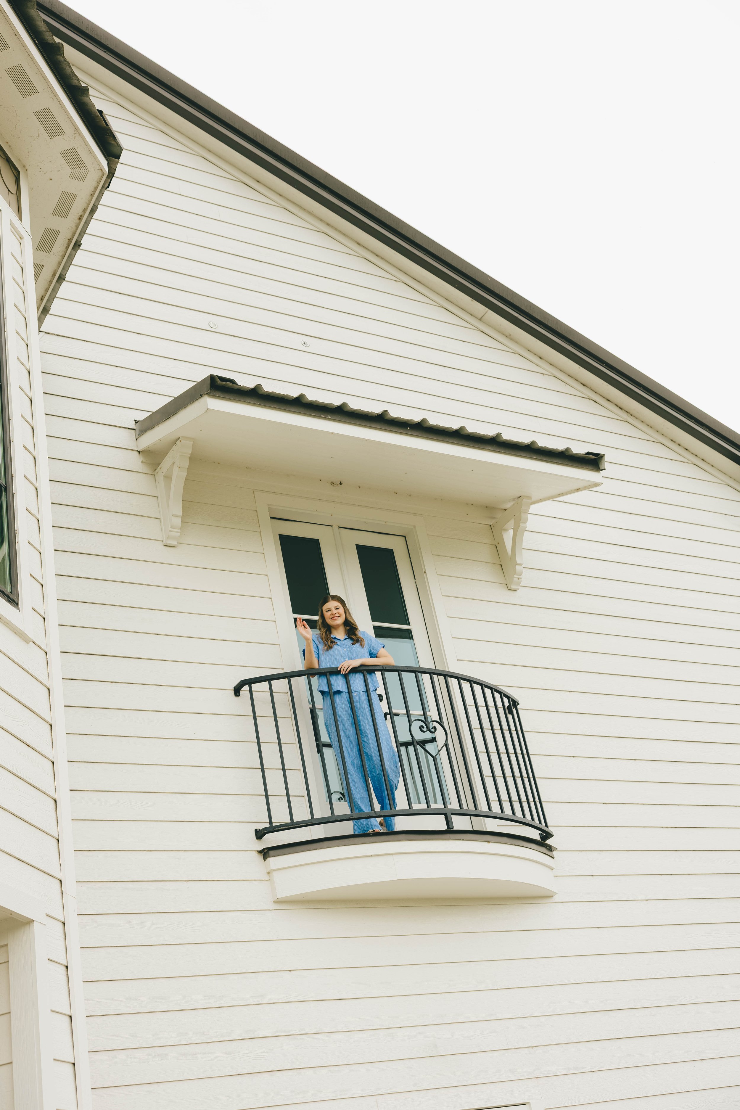 A woman standing on a small balcony of a white house with horizontal siding, smiling and waving with her right hand, dressed in a loose blue top and matching blue pants.