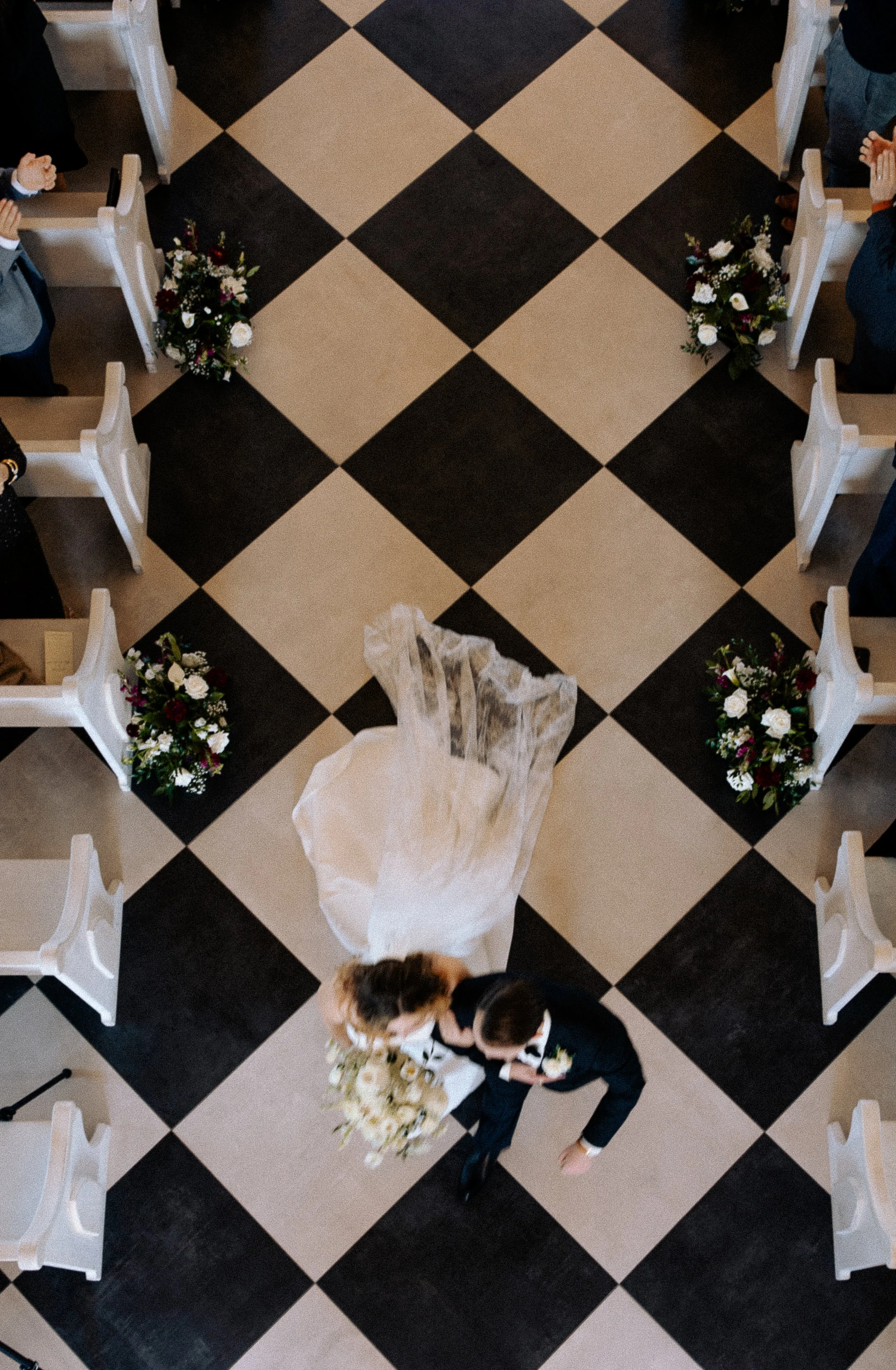 Top-down view of a bride and groom walking down the aisle in a wedding ceremony, surrounded by white chairs and floral arrangements on a black and white checkered floor.