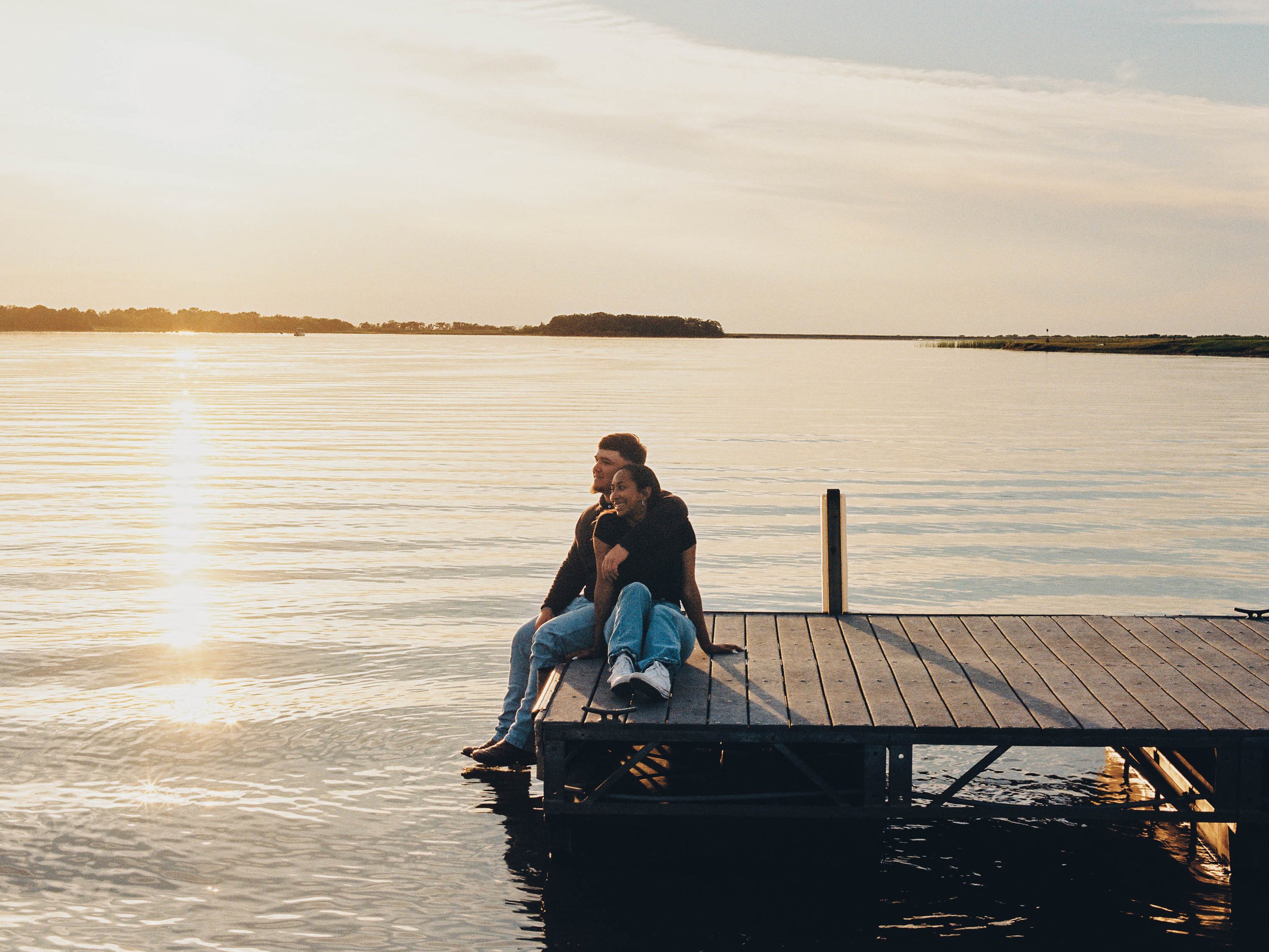 A couple sitting on a dock by the water during sunset, with the sun reflecting on the water surface and nature in the background.