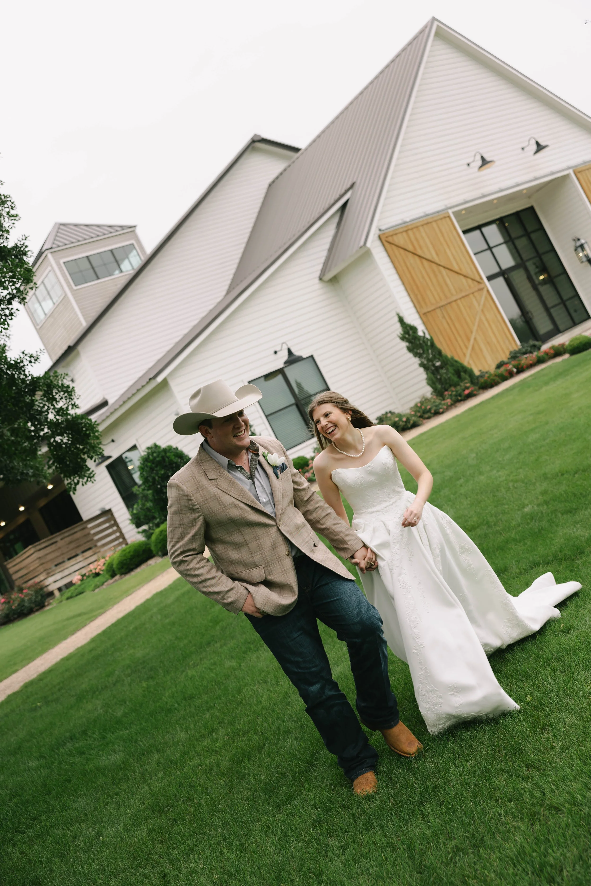A bride and groom walking hand in hand on a grassy lawn outside a modern white house with wooden accents, smiling and enjoying their wedding day.