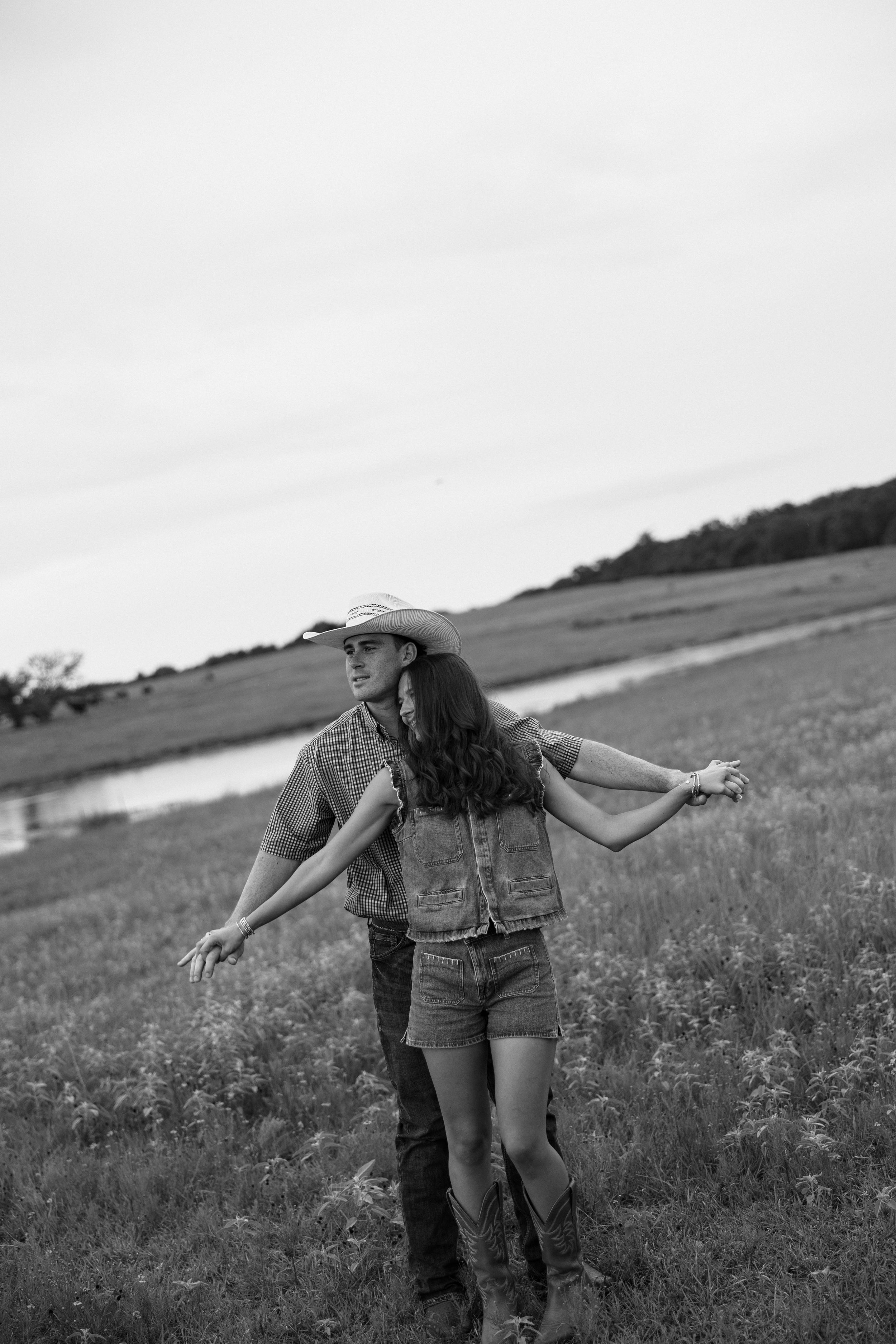Young man in a cowboy hat and young woman in jeans and a sleeveless denim vest standing in a field, dancing with arms outstretched.