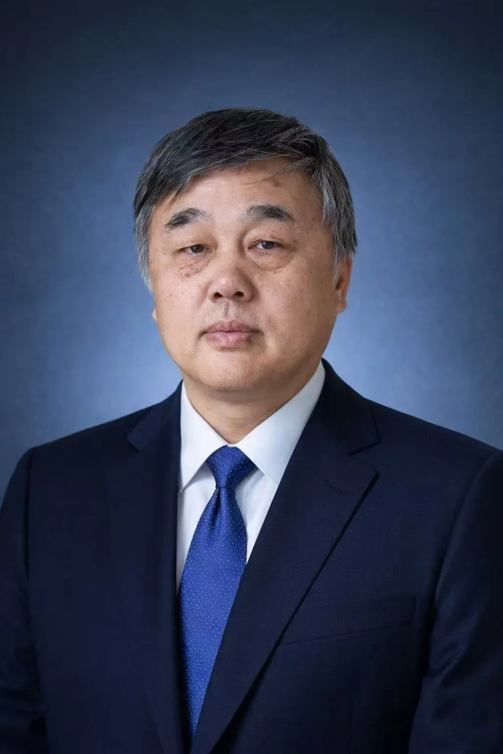 A professional portrait of an East Asian man in a dark suit, white shirt, and blue tie, with a neutral background.