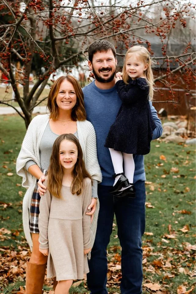 A family of four smiling outdoors on fall day, standing in front of a tree with red berries. The mother has shoulder-length red hair, wearing a white cardigan and patterned skirt. The father has dark hair and beard, wearing a blue shirt. The young girl in beige dress stands in front, and the other girl in black dress is being held by her dad.