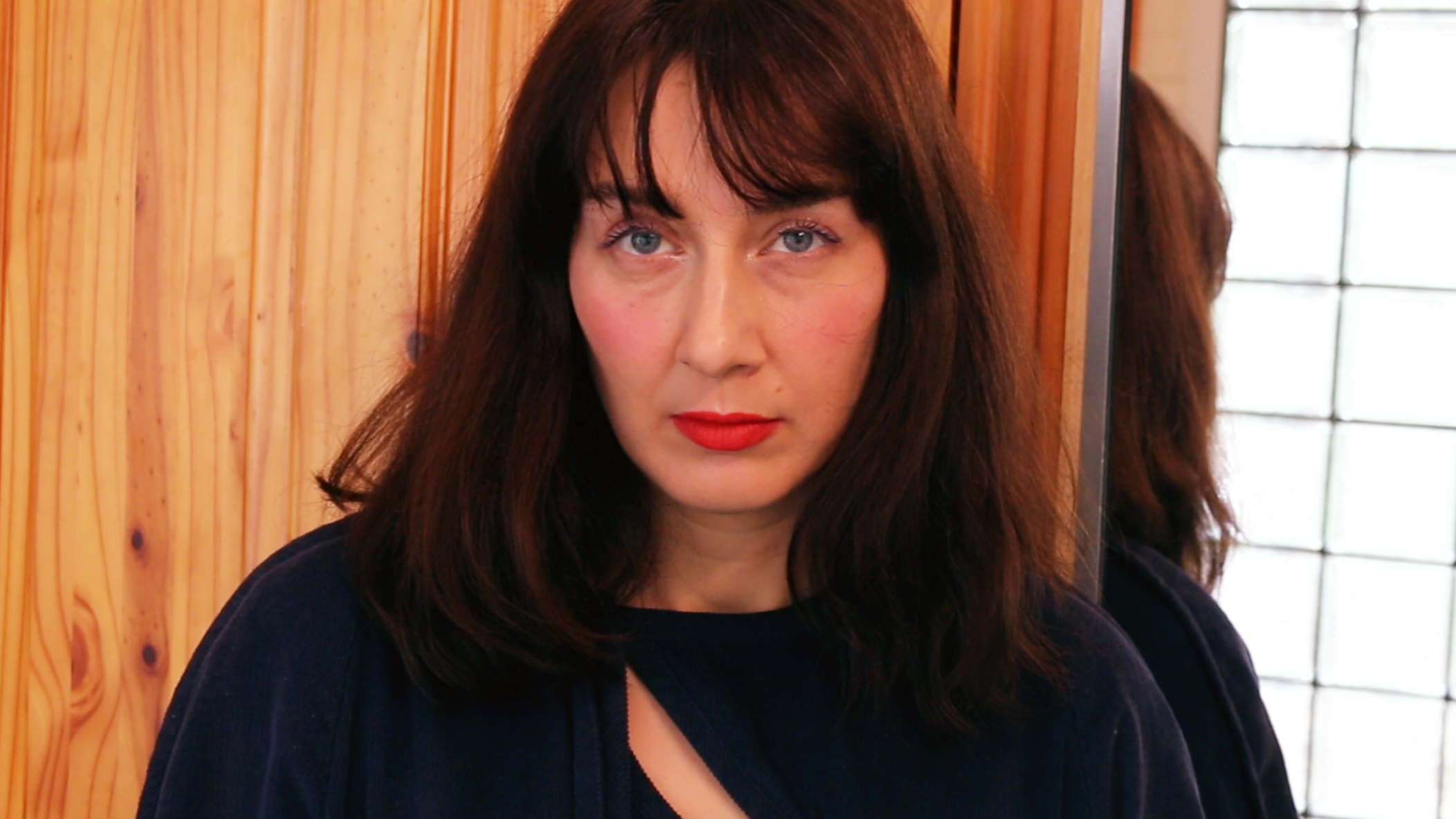 A woman with long brown hair, blue eyes, and red lipstick standing in front of a wooden wall and mirror.