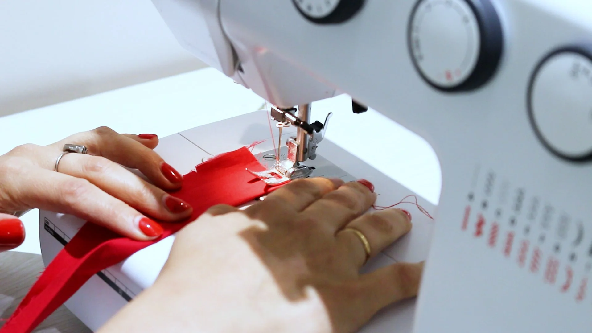 Close-up of hands sewing red fabric on a white sewing machine, with visible control dials and a piece of paper on the side.