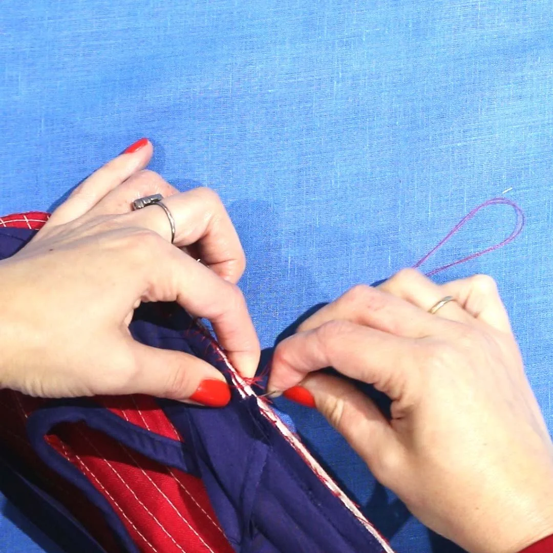 Close-up of hands sewing red and navy fabric with red thread doing a whip stitch, on a blue surface.