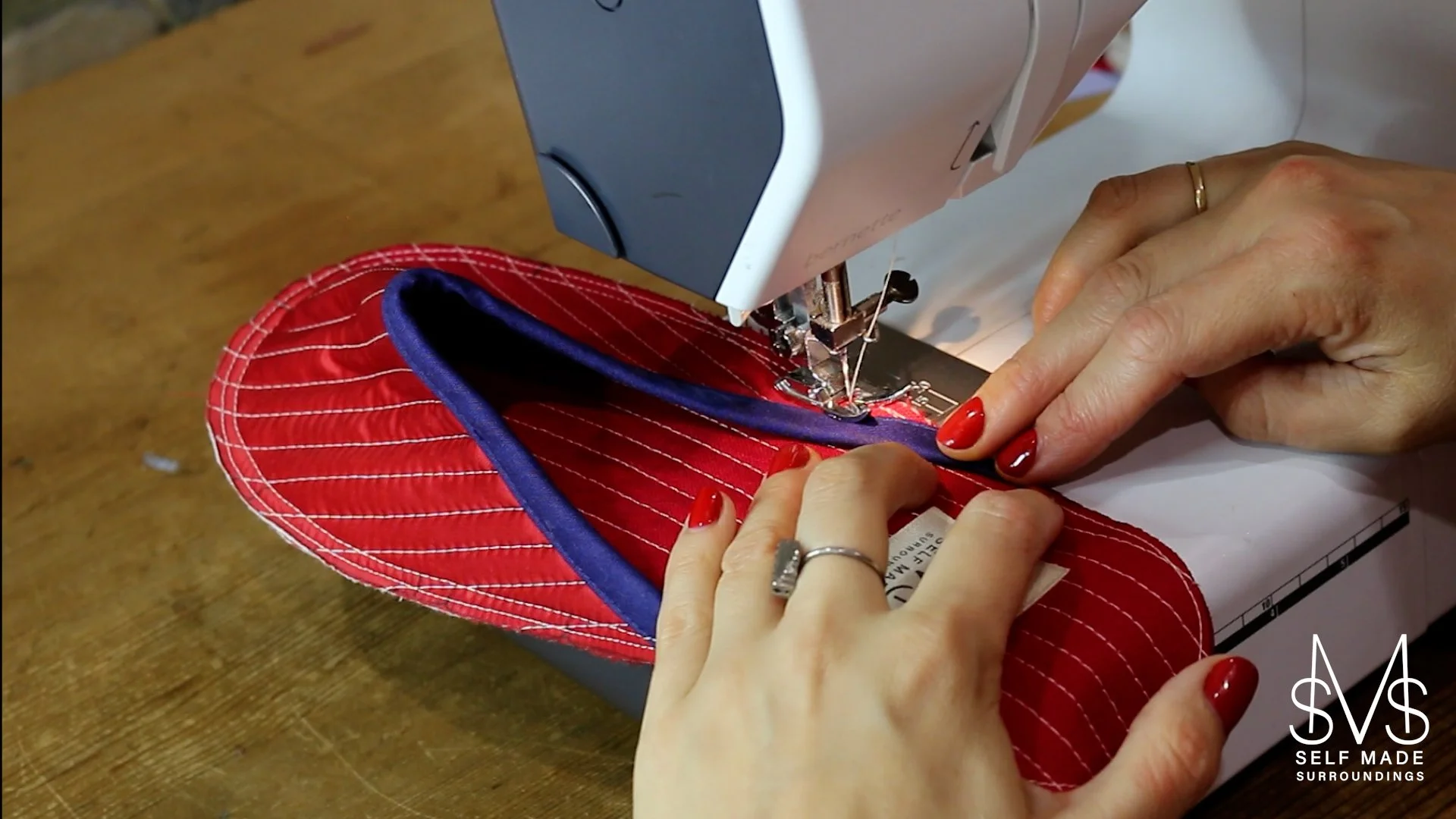 A person sewing a red quilted fabric slipper with navy bias edging on a sewing machine, with a wooden table in the background.