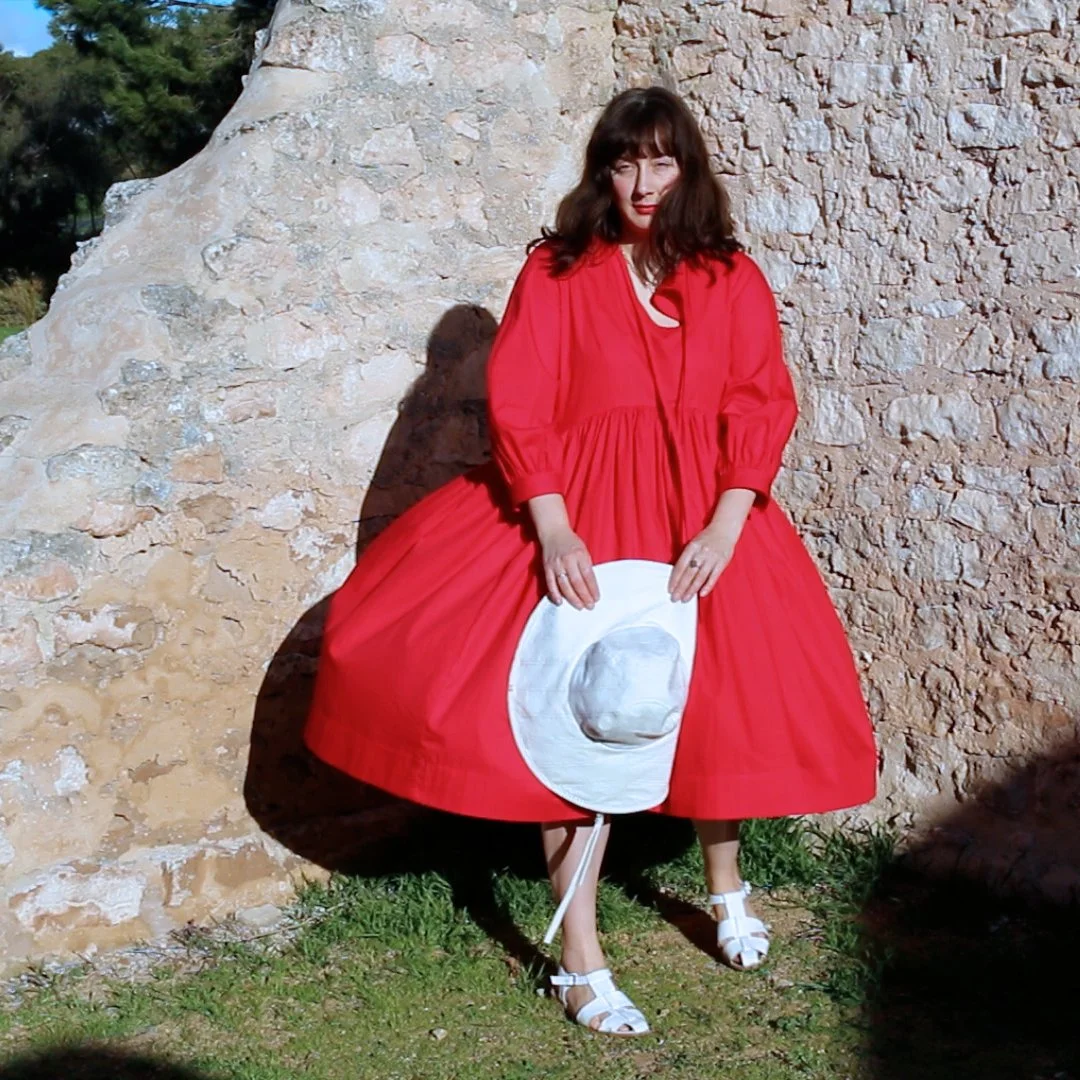 A woman wearing a bright red dress and white sandals is standing outdoors against a stone wall. She is holding a white hat with a circular design and has dark brown, wavy hair.