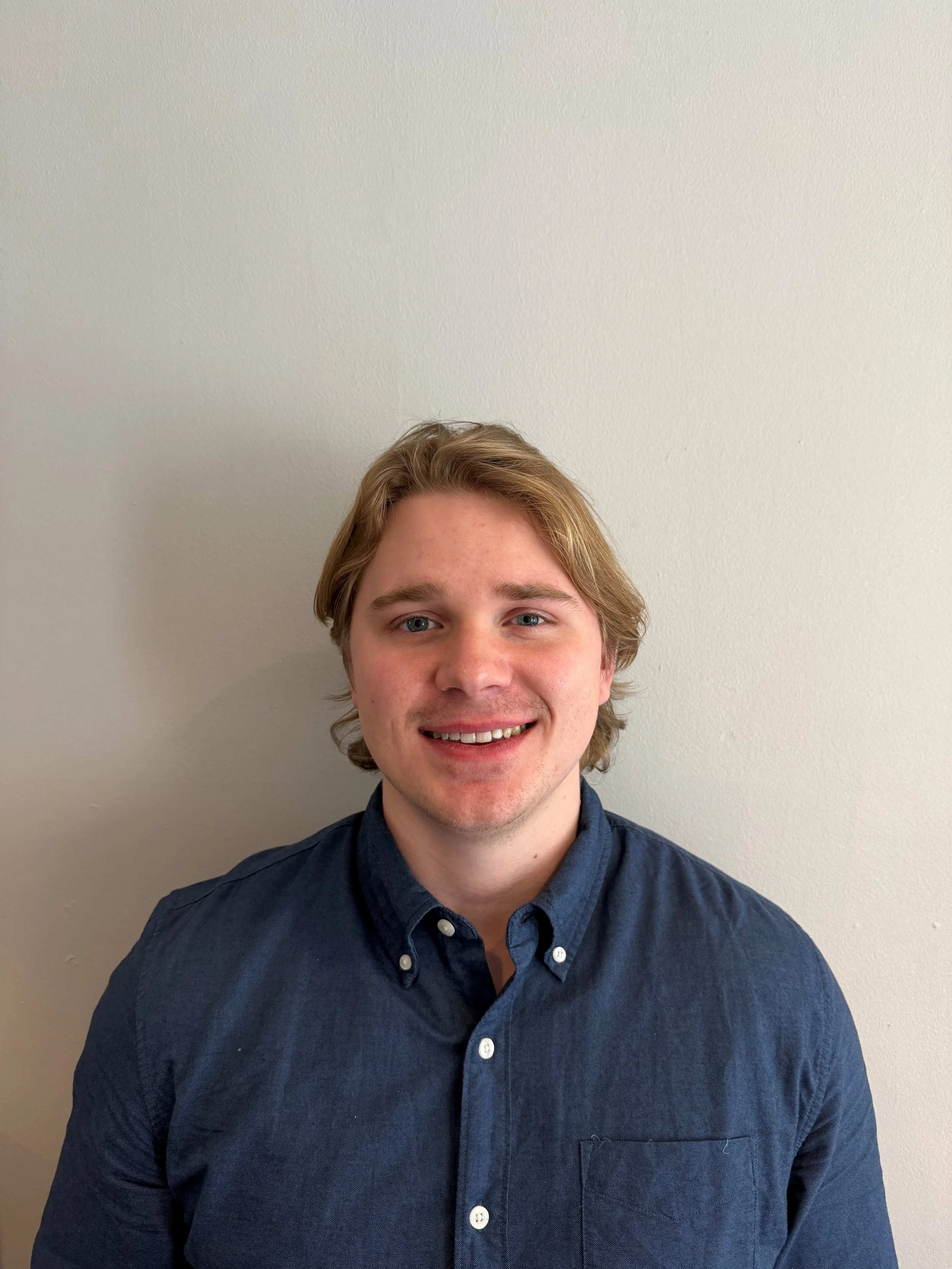A young man with blonde hair and blue eyes smiling, wearing a dark blue button-up shirt, standing against a plain gray wall.