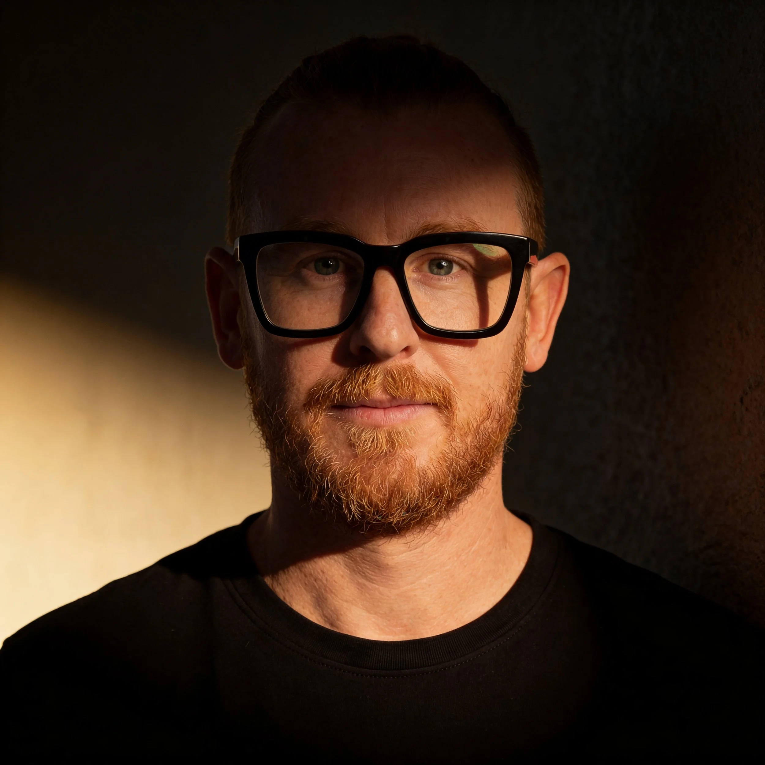 Portrait of a man with red hair and beard wearing large black glasses, against a dark background.
