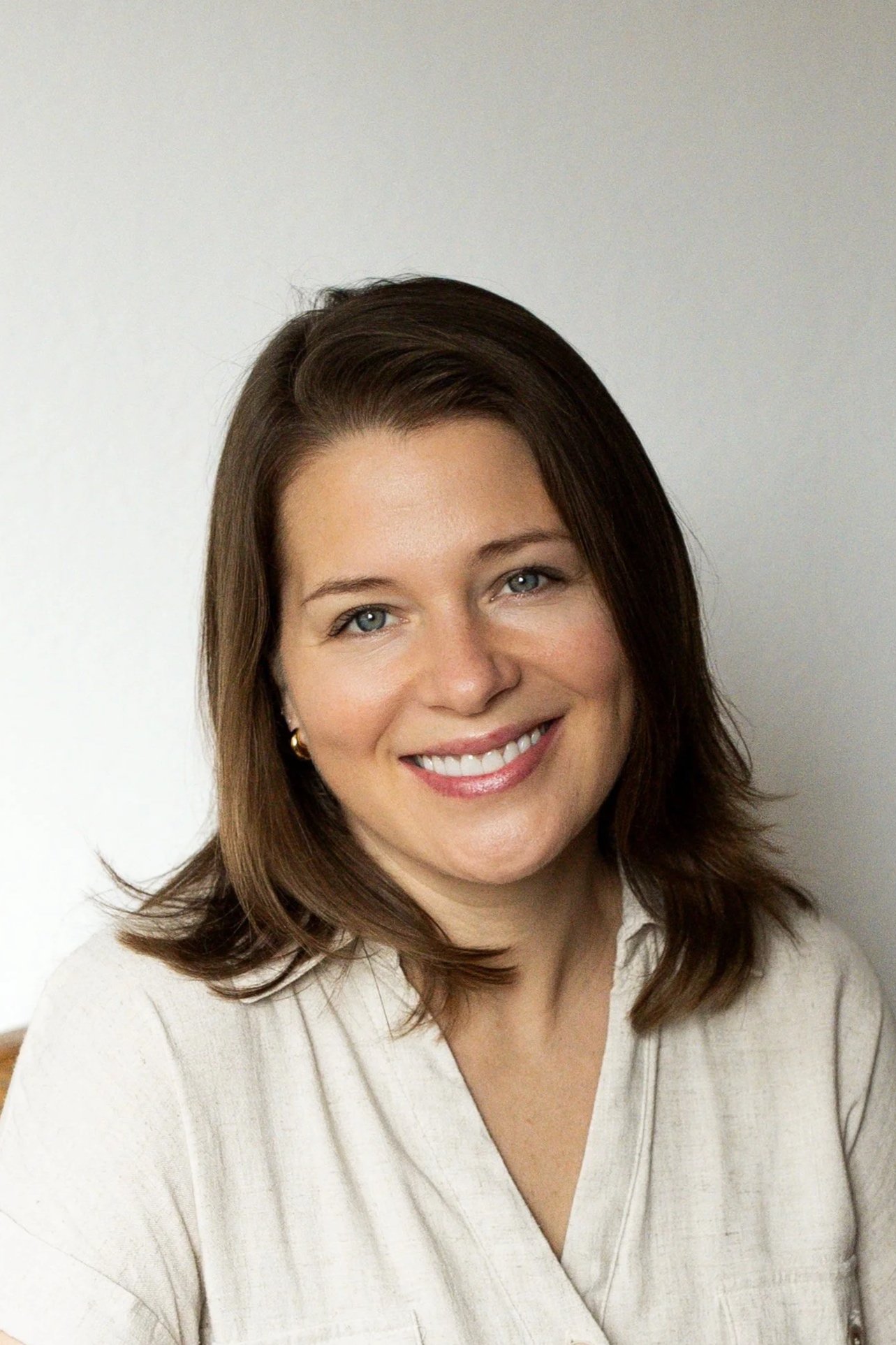 A woman with shoulder-length brown hair, blue eyes, and a bright smile wearing a light beige top against a plain white wall.