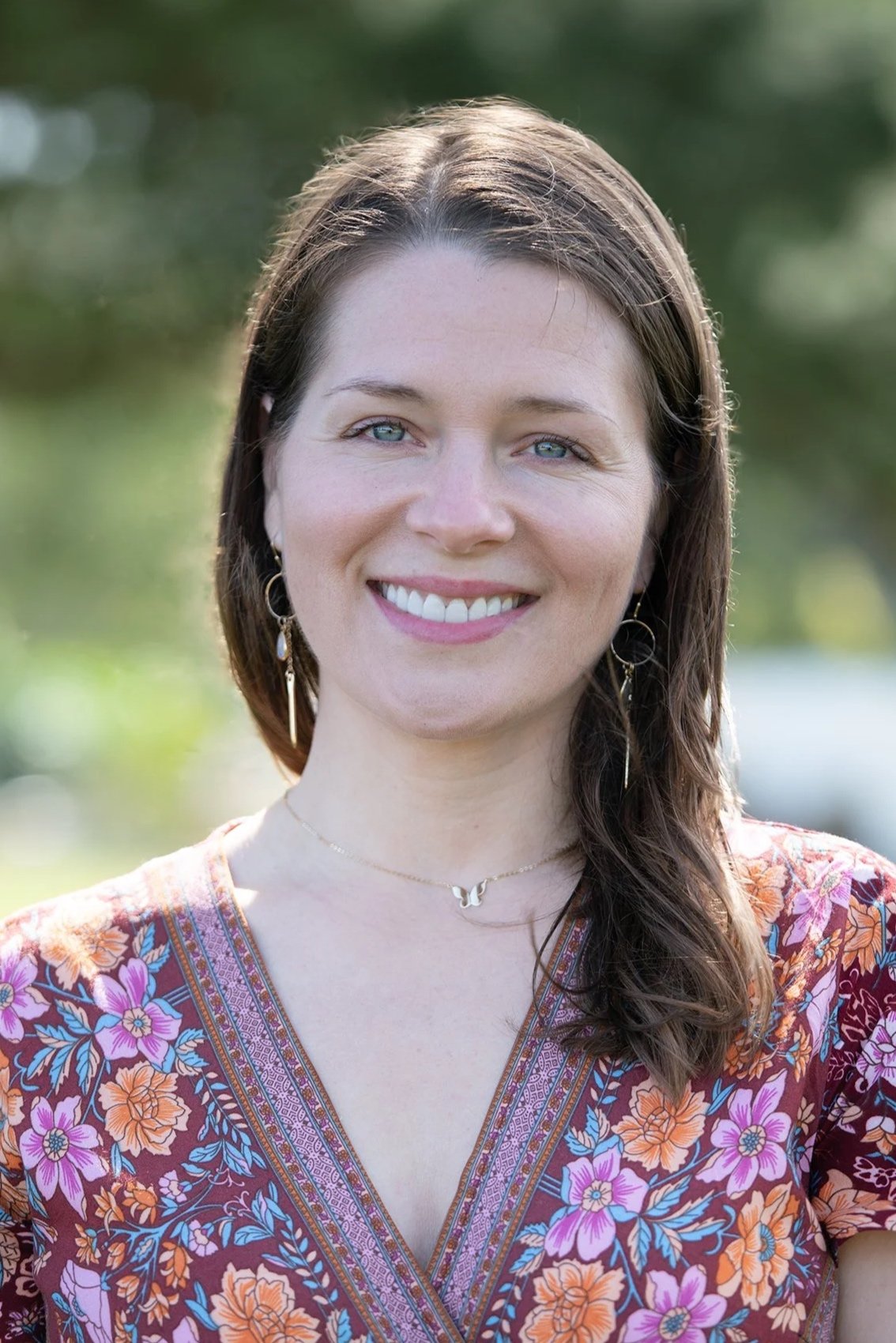 A woman with long dark hair, blue eyes, and light skin smiling outdoors, wearing a colorful floral top and jewelry.
