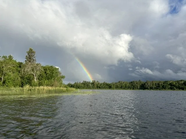 A lake scene with trees along the shoreline and a rainbow in the cloudy sky.