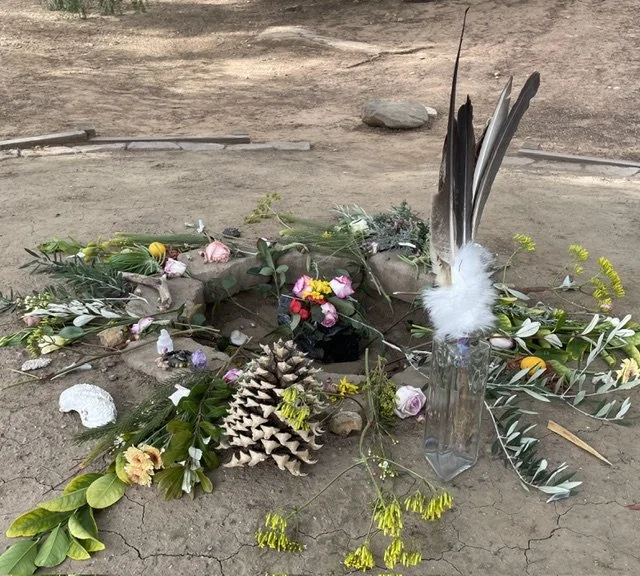 A makeshift outdoor memorial with flowers, pine cone, feathers in a glass jar, and other natural objects arranged on the ground.