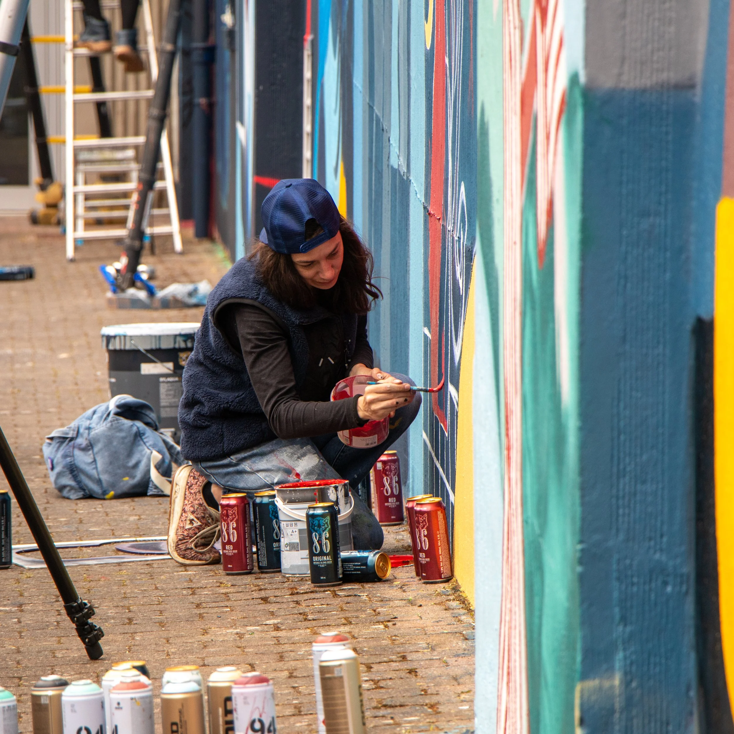 Une femme en train de peindre une fresque murale colorée à l'extérieur, avec des bombes de peinture à ses pieds et une échelle à proximité. PORTRAIT OF LYSEOPAINE