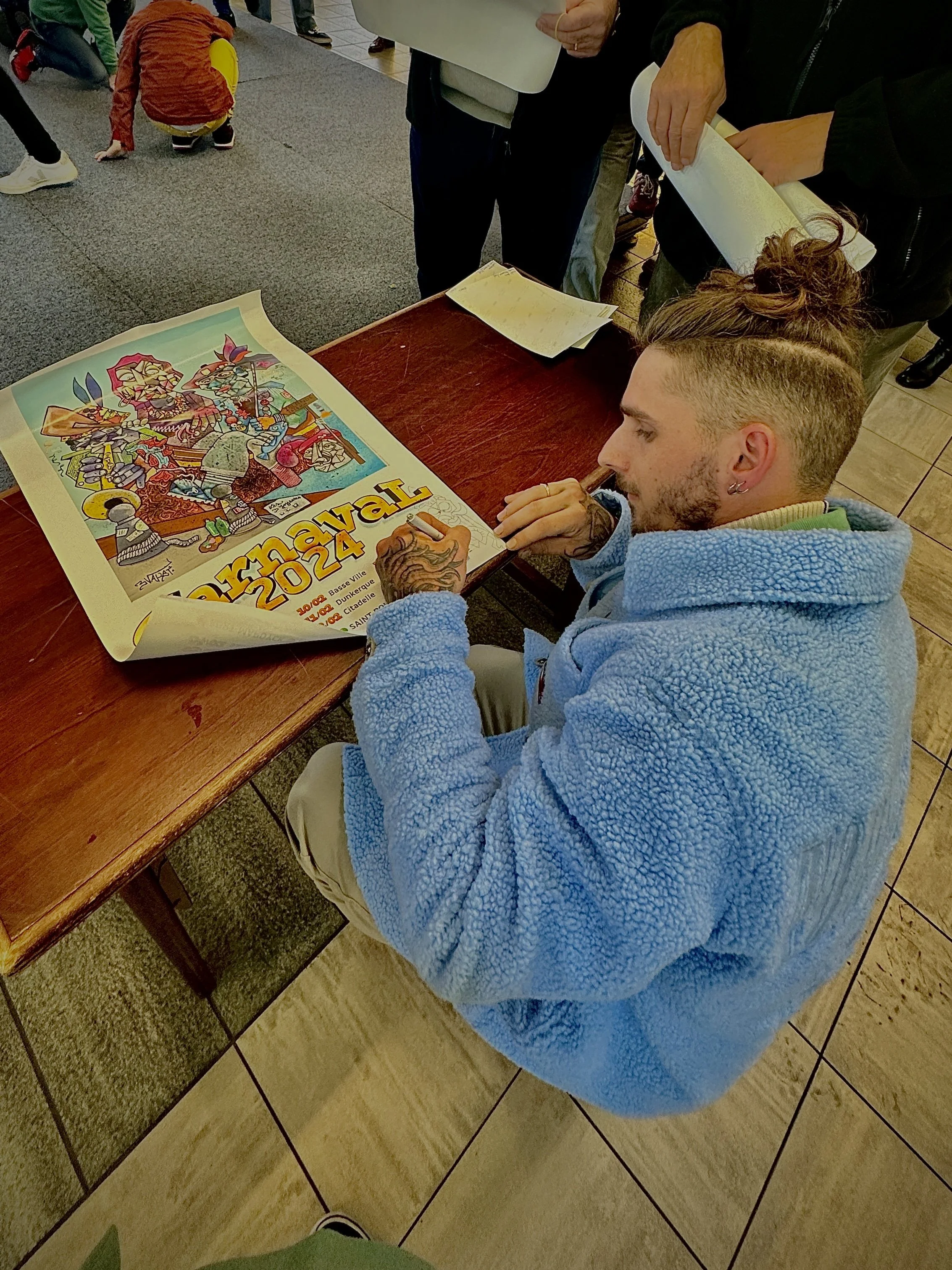 Un homme avec des tatouages dans un hoodie bleu, assis à une table, signing ou faisant une dédicace à un poster coloré de 2022, à côté de plusieurs personnes en intérieur.