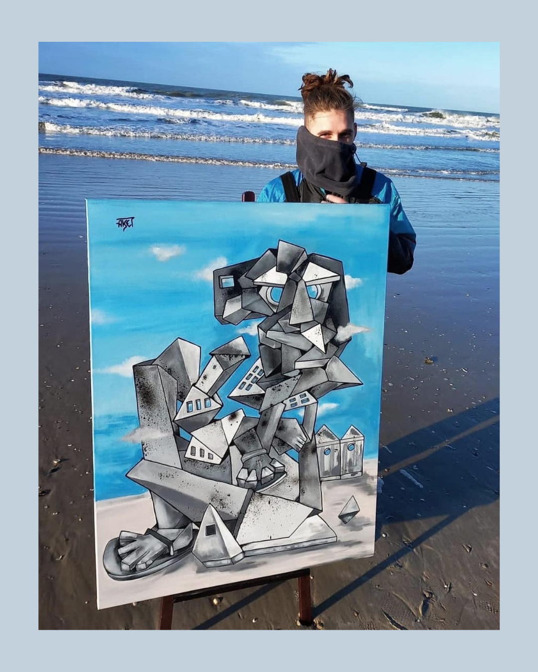 Une jeune femme debout sur la plage tient une peinture représentant un lion stylisé en style graffiti, avec un fond de ciel bleu avec quelques nuages.